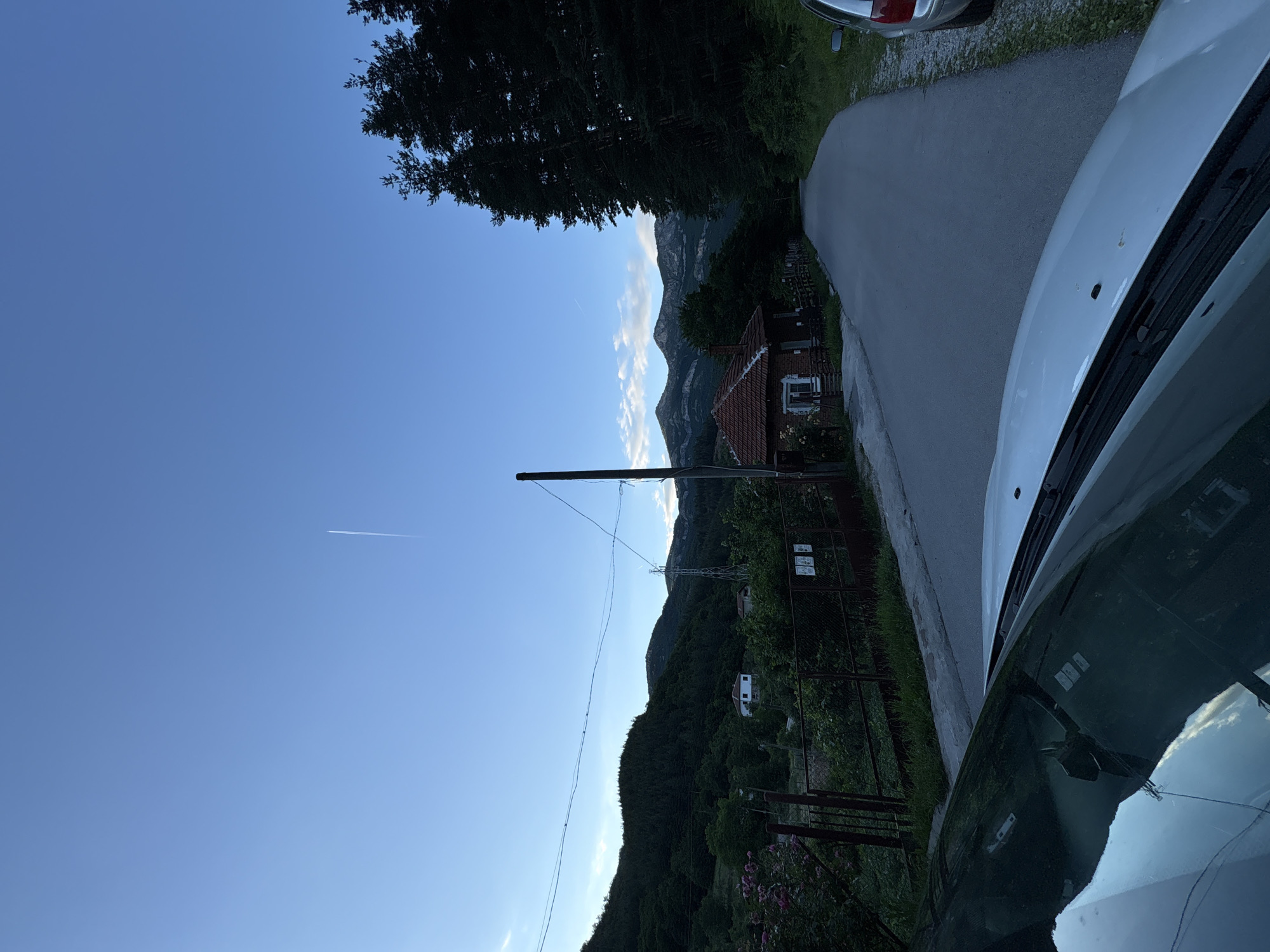 The image depicts a rural road in the city of Zgorigrad, Bulgaria, at dusk. A paved road curves through a village setting. On the left side of the road, a small, red-tiled roof house with a brick exterior is visible, nestled among trees and a fence. Further up the hill on the left, a larger white house with a red roof can be seen. The background is dominated by lush, dark green forested hills and mountains under a clear blue sky with a faint contrail from an airplane. On the right side of the road, tall, dark green pine trees line the landscape. A silver car is parked on the right side of the road. The foreground shows the hood and windshield of a white car, reflecting the sky and surroundings. A single utility pole stands near the house on the left.