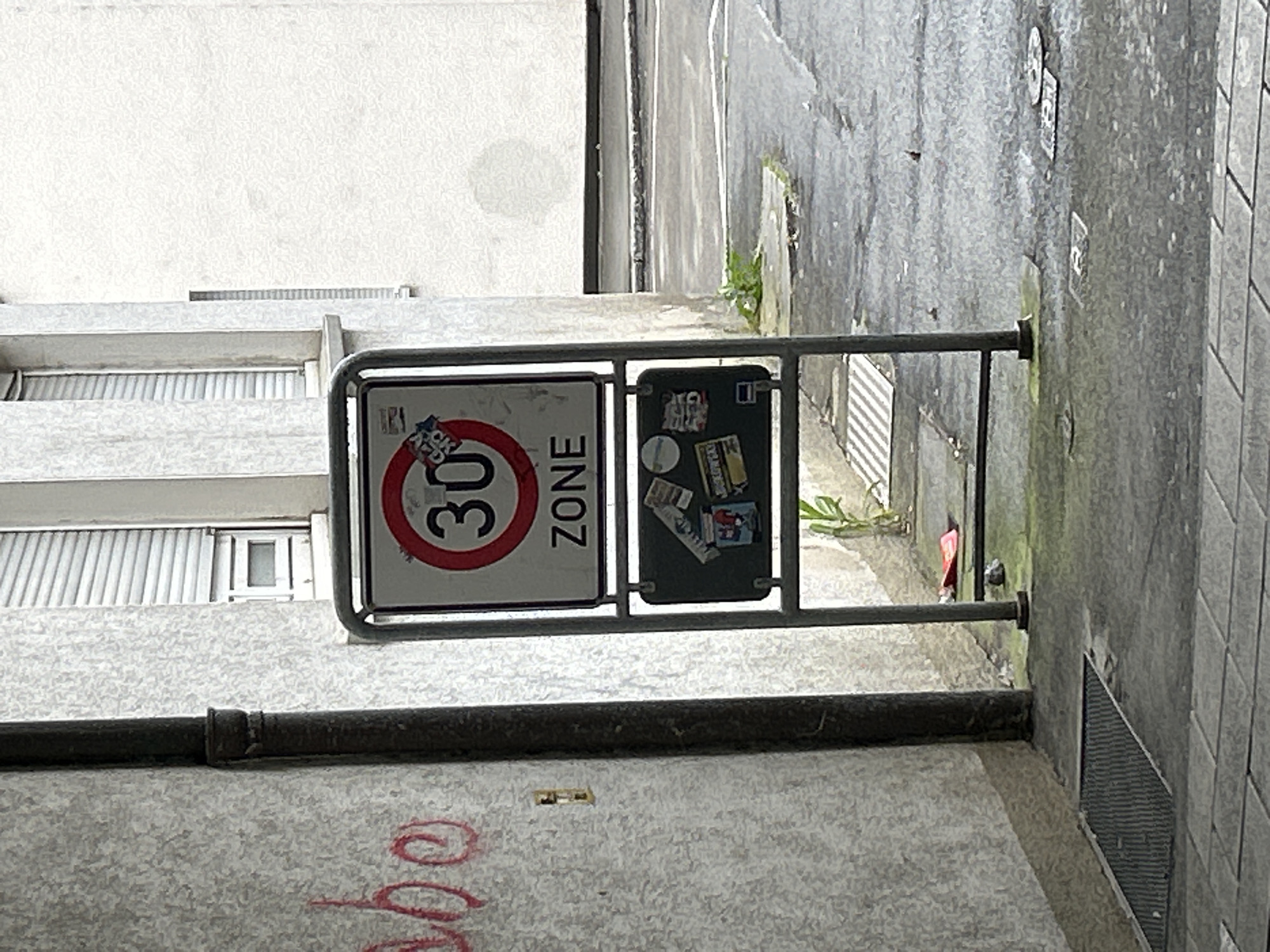 A close-up, rotated photograph shows a speed limit sign displaying "30 ZONE" in a red circle, attached to a gray metal frame. A second sign with stickers is also attached to the frame. The background includes a light-colored building with closed blinds, an alleyway with concrete walls, and a drain. Graffiti is visible on the lower portion of the image. The location is Unknown, Unknown.