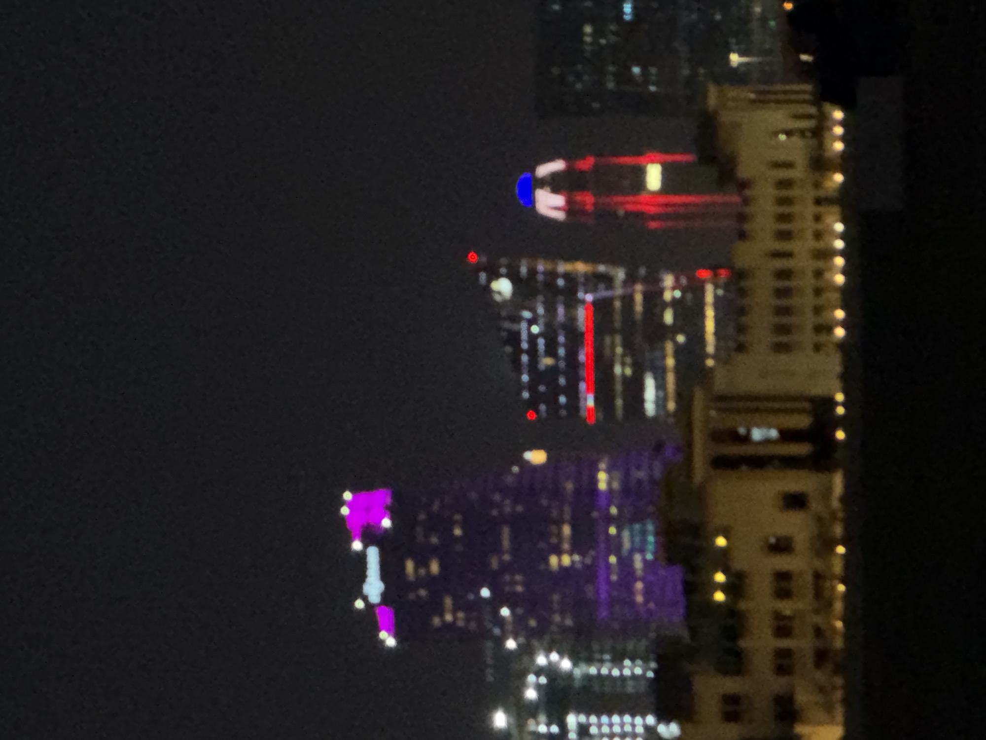 The image captures a nighttime cityscape with several illuminated buildings. The prominent structure on the left is a skyscraper with vibrant purple lighting covering its upper sections and some horizontal stripes. To its right, another building is visible, featuring red horizontal lighting and a red circular light at its apex, with a blue dome-like structure above it. A third, more distant skyscraper shows white and red lights. In the foreground, a low-rise building with illuminated windows and decorative arches is visible, appearing to be a historical or classical architectural style. The overall scene is dark, with the city lights serving as the primary illumination. The image is slightly blurry, suggesting it was taken from a distance or with a handheld camera.