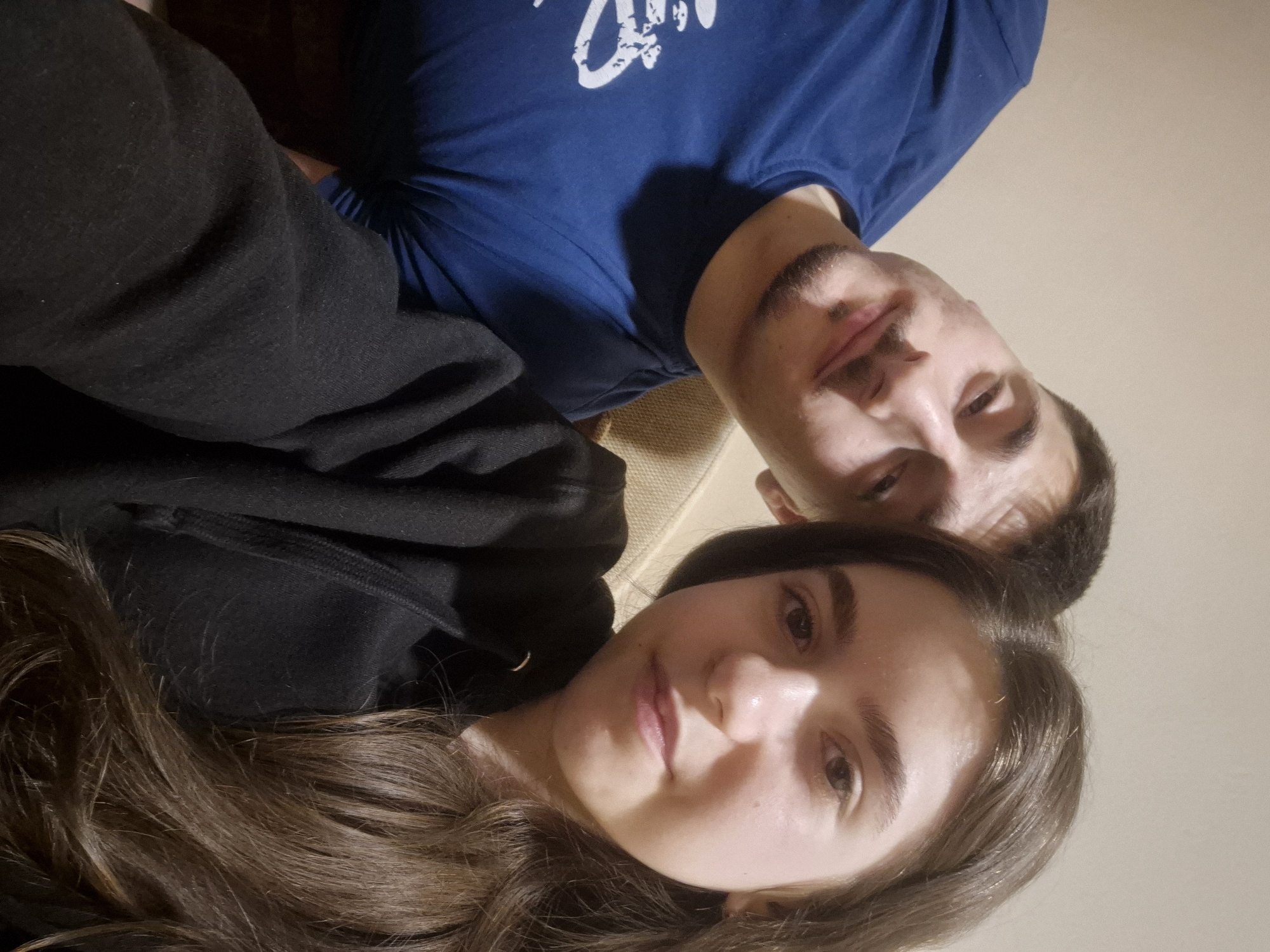 A selfie shows a man and a woman indoors. The man is wearing a blue t-shirt with a white design on it. He has dark hair and a mustache. The woman has long brown hair and is wearing a black hooded sweatshirt. The background is a plain light color. There is no information about the location in the city of Pavlikeni, Bulgaria, visible in the image.