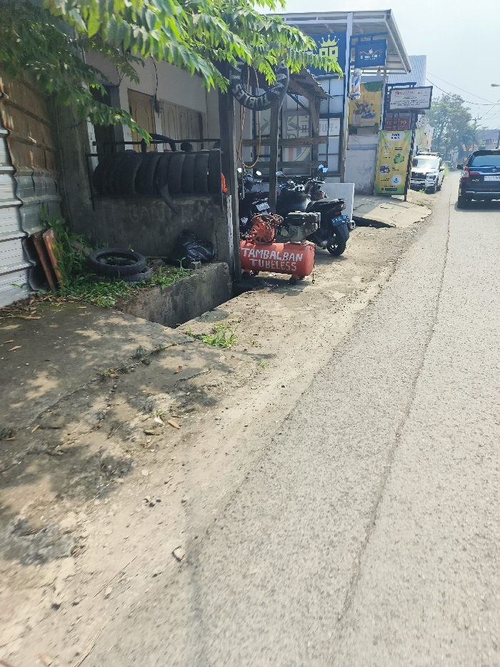 The image shows a street in Cileunyi, Indonesia, at the coordinates -6.946 latitude and 107.725 longitude. The street is paved and has a sidewalk on the left side. There is a small, open-air shop on the left side of the street, with several tires stacked against the wall. There is a small ditch in front of the shop, and some green plants are growing in the ditch. There are two motorcycles parked in front of the shop, and a red, rectangular object with the words "Tambalban Tubeless" on it is sitting in front of them. The shop appears to be closed. There are two cars parked on the opposite side of the street, with a few cars passing by. The street is relatively empty, with no people visible. The sky is clear and blue.