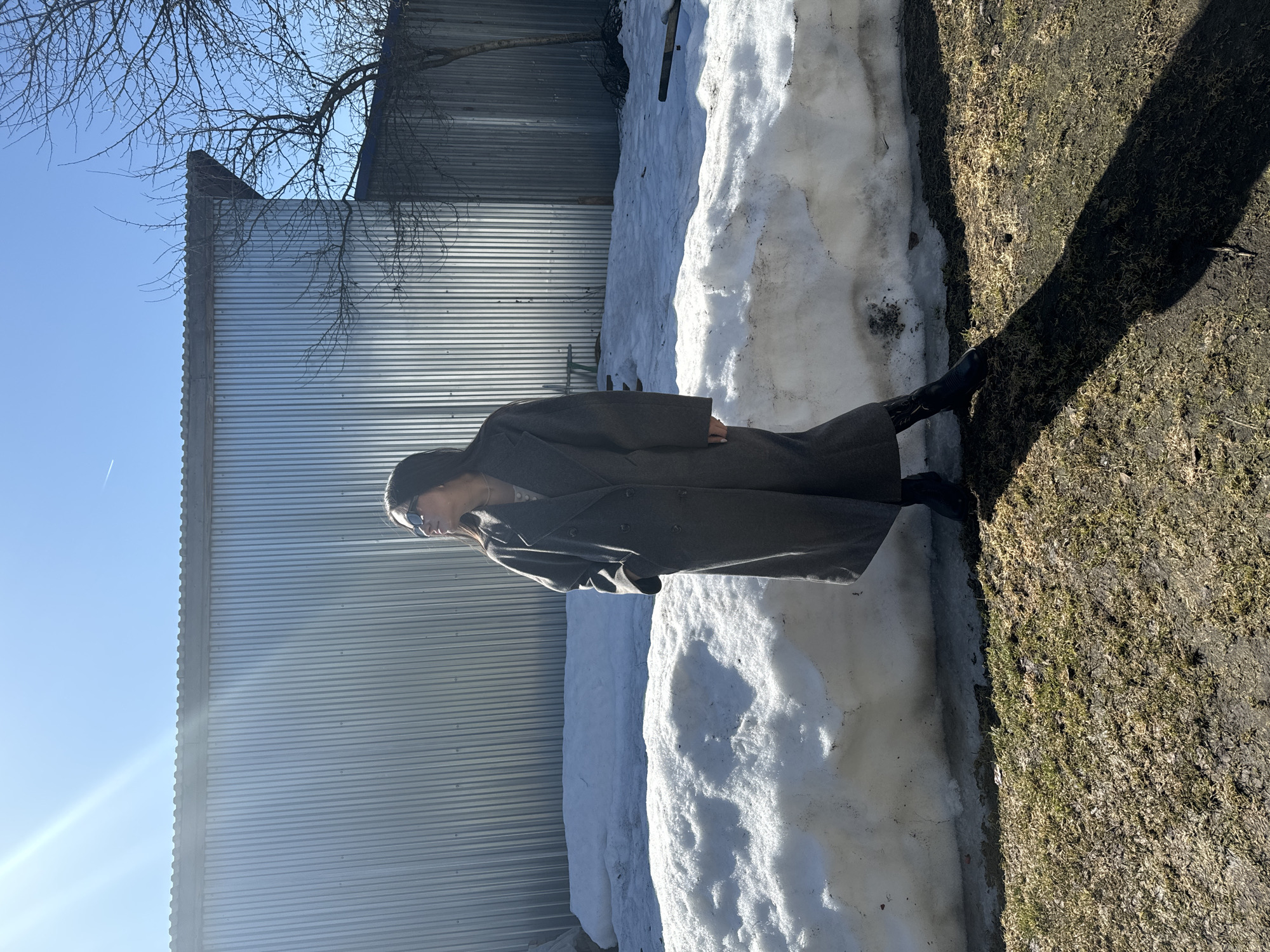 A woman wearing a dark overcoat and sunglasses stands next to a snowdrift. The location appears to be outdoors, adjacent to a metal corrugated building or fence. The ground consists of patches of grass and bare earth. The sky is visible and blue. It is impossible to forensically identify the location as Красный Маяк, Russia, but it is wintertime.