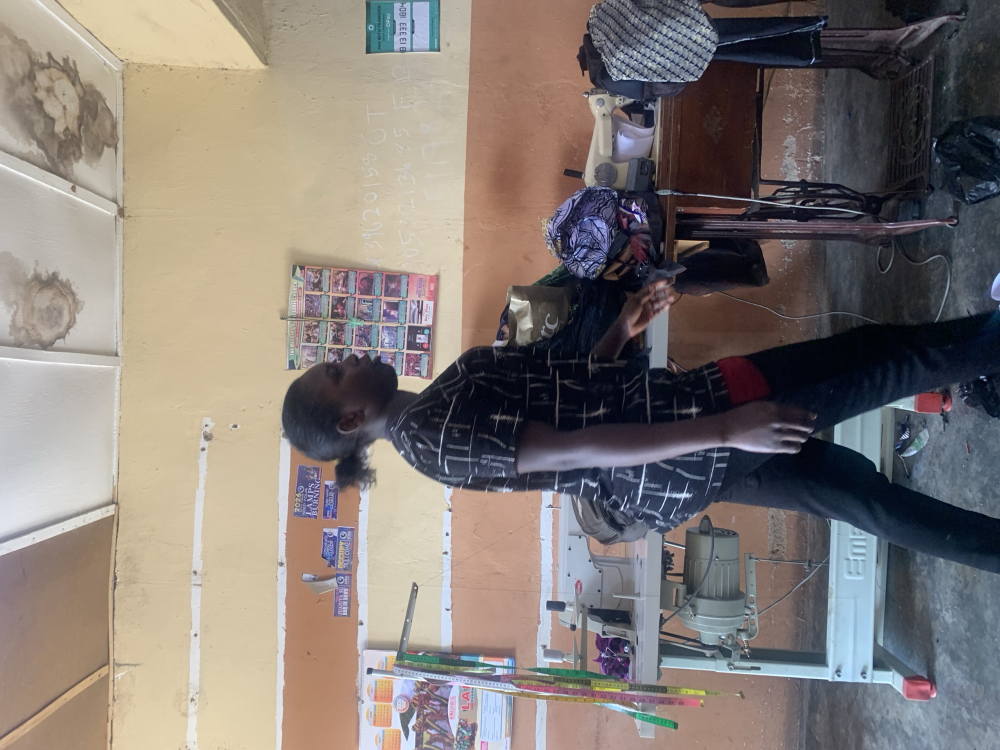 A full-shot, medium-distance, eye-level, side profile, outdoor, daytime photograph taken in Jos, Nigeria shows a Black female tailor walking from left to right past a sewing machine. The woman is wearing a black shirt with white lines and black pants, and her hair is pulled back into a bun. She has a red object in her right hand. The scene appears to be a tailor shop, with sewing machines, fabric, and measuring tapes visible. The walls of the shop are a light yellow color with orange accents. The ceiling has water stains.