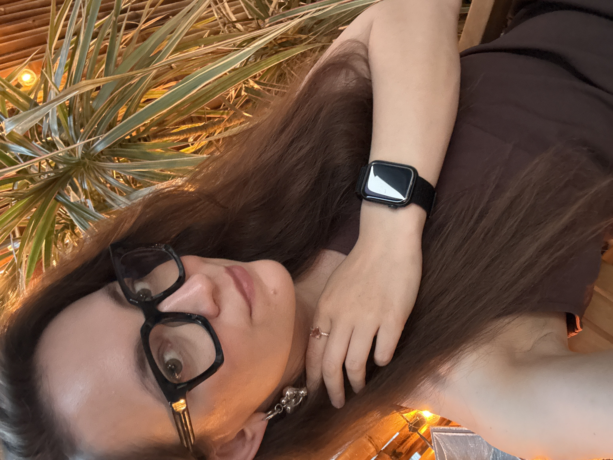 A woman with brown hair, wearing black-rimmed glasses, a brown dress, and an Apple Watch with a black band, is lying down. A ring is on her finger, and dangling earrings are visible. Plant leaves are in the background. The location cannot be determined from the image.
