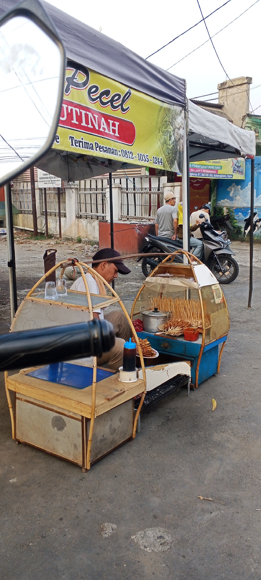 An outdoor scene depicts a street food vendor's stall, specifically for "Pecel" (a Javanese salad with peanut sauce), in Depok, Indonesia. The stall features a yellow banner with red lettering that reads "Pecel JUTINAH" and "Terima Pesanan: 0812-1035-1244". Two men are present. One man, wearing a black cap and white shirt, is seated behind a food cart, possibly preparing or serving food. Another man, wearing a beige cap and grey t-shirt, is seated on a motorcycle parked behind the stall. A third man, wearing a white t-shirt and a beige cap, is also seated on a motorcycle. The food cart itself is made of bamboo and wood, with a transparent canopy displaying skewers of food, a pot, and a bottle of sauce. In the foreground, a motorcycle's side mirror is visible, reflecting the sky and part of the food stall's banner. The environment appears to be a roadside setting with a paved surface, a fence in the background, and some debris on the ground. The general atmosphere suggests a commercial or service area within the city.