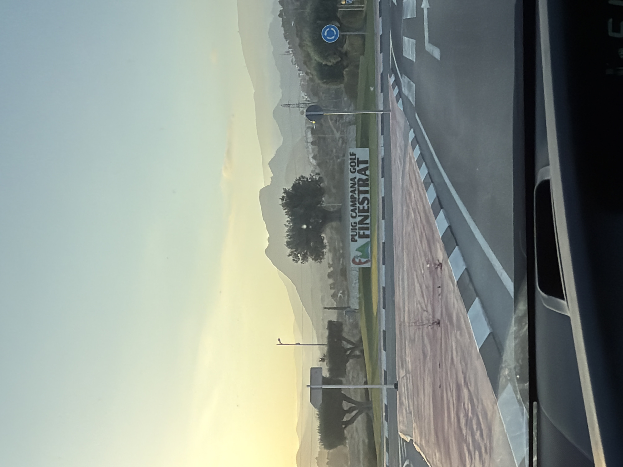 The image captures an early morning view in Finestrat, Spain, with the sun low on the horizon, casting a warm, yellow glow across a hazy landscape of rolling hills and distant mountains. In the foreground, a road curves to the right, marked with white and grey traffic calming features and pedestrian crossings. To the left of the road, a patch of reddish-brown ground is visible, interspersed with small, sparse trees.
Further back, a white sign with green accents reads "PUIG CAMPANA GOLF FINESTRAT". The sign is situated behind a low, green-trimmed embankment and in front of a line of mature, manicured trees. A solitary lamppost stands near the sign, and beyond that, more trees and a gentle slope lead up to the hazy, layered mountains that dominate the background. The sky is a soft gradient of pale yellow near the horizon, transitioning to a light blue. No people are visible in the scene. The overall impression is one of a peaceful, early morning landscape at a golf resort.