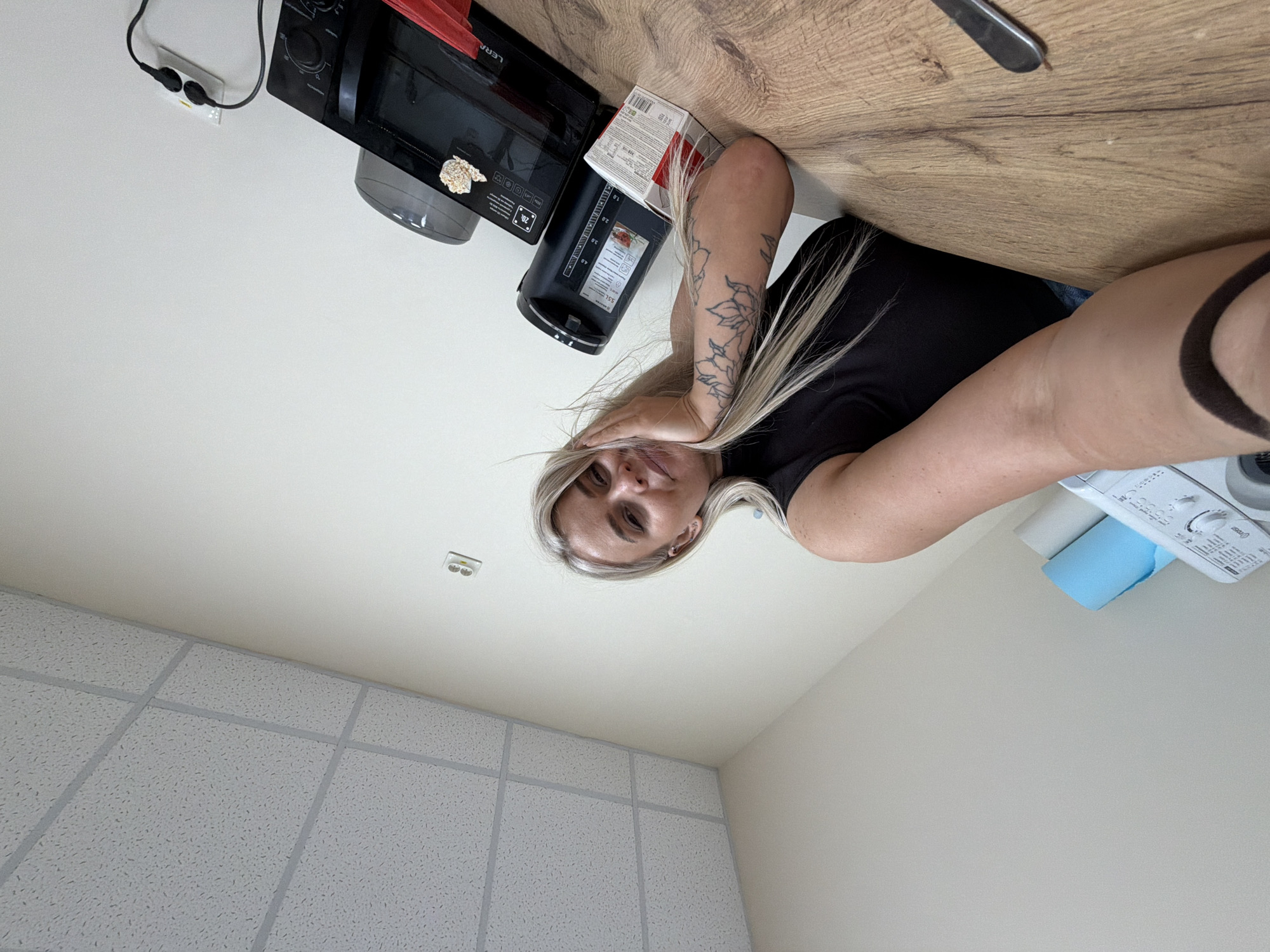 A woman with blonde hair, wearing a black top and jeans, is visible in the image. Her left arm has a floral tattoo. She's indoors, resting her head on her left hand. A microwave oven, kettle, and a box are on the table next to her. A washing machine and a roll of blue paper towels are also visible in the background. The ceiling is white with a grid pattern. The location is not determinable as Sarapul, Russia from this image alone.