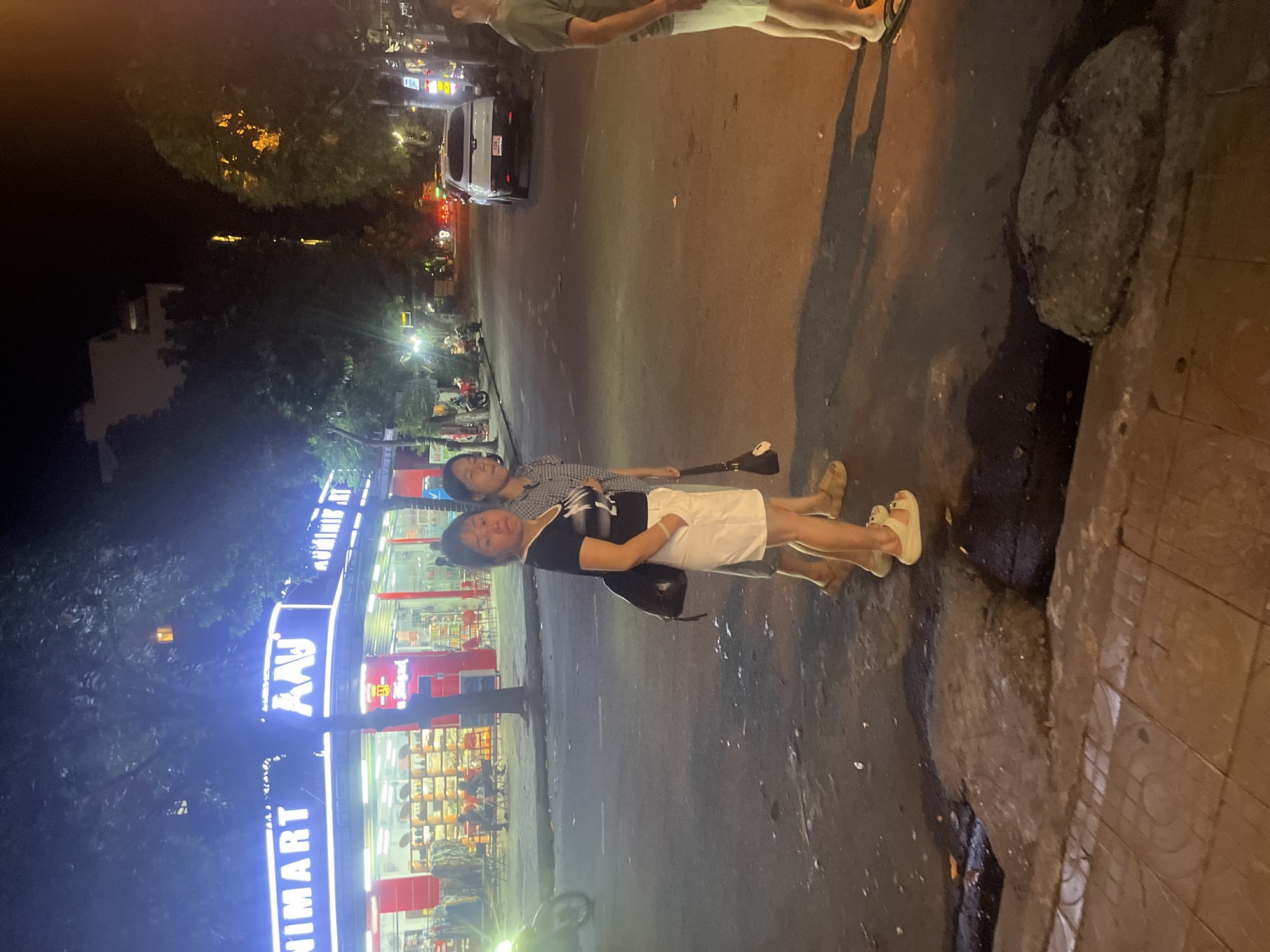 The image shows a street scene at night in Da Nang, Vietnam. In the foreground, two women are walking on the sidewalk. The woman on the left is wearing a black t-shirt with a graphic print and white shorts. She is carrying a black bag over her shoulder. The woman on the right is wearing a plaid shirt and is also walking. Behind them, a white car is parked on the street. To the left of the women, there is a brightly lit store with "NIMART" and "AAW" written in neon letters. The store appears to be a supermarket or convenience store, with shelves visible inside. Trees line the street, and streetlights illuminate the area. The overall atmosphere is that of a busy urban environment at night.