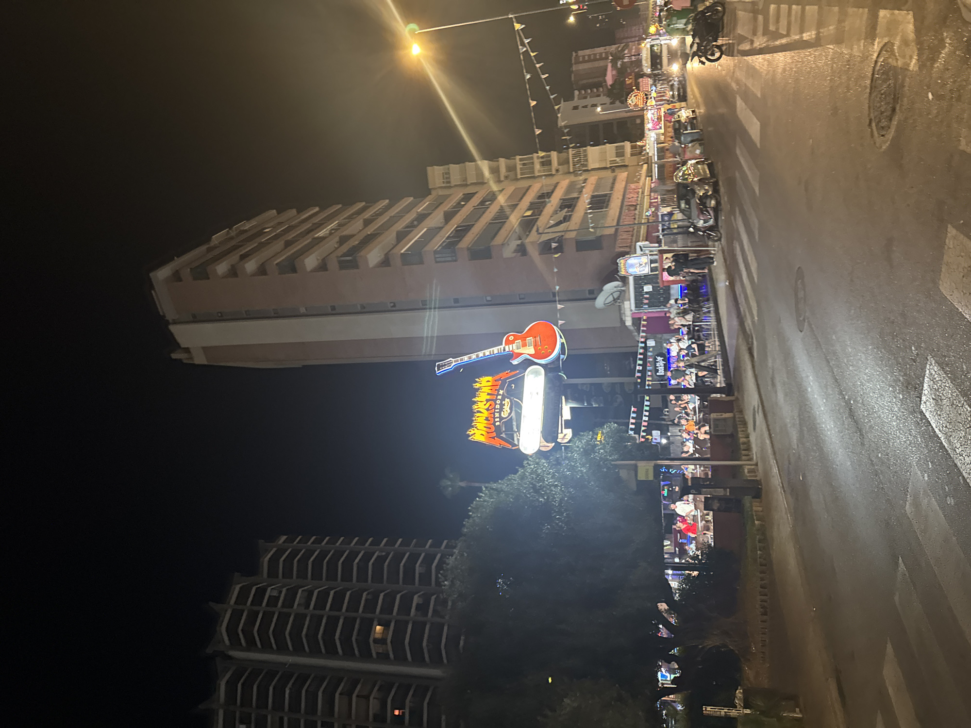 The image captures a nighttime scene on a street in Benidorm, Spain. Dominating the left side of the frame are two tall, multi-story buildings with numerous balconies, characteristic of the city's architecture. In the center, a prominent neon sign in the shape of a guitar, with flames emanating from the top and the word "ROCKSTAR" in bold lettering, is illuminated. Below this sign, a restaurant or bar area is visible with outdoor seating and people gathered. The street is wet, suggesting recent rain, and reflects the bright lights of the establishments. Several cars and a motorcycle are parked or stopped on the street, and pedestrian crossings are marked with white lines. Decorative string lights are strung across the street in some sections. The overall atmosphere is that of a lively entertainment district at night.