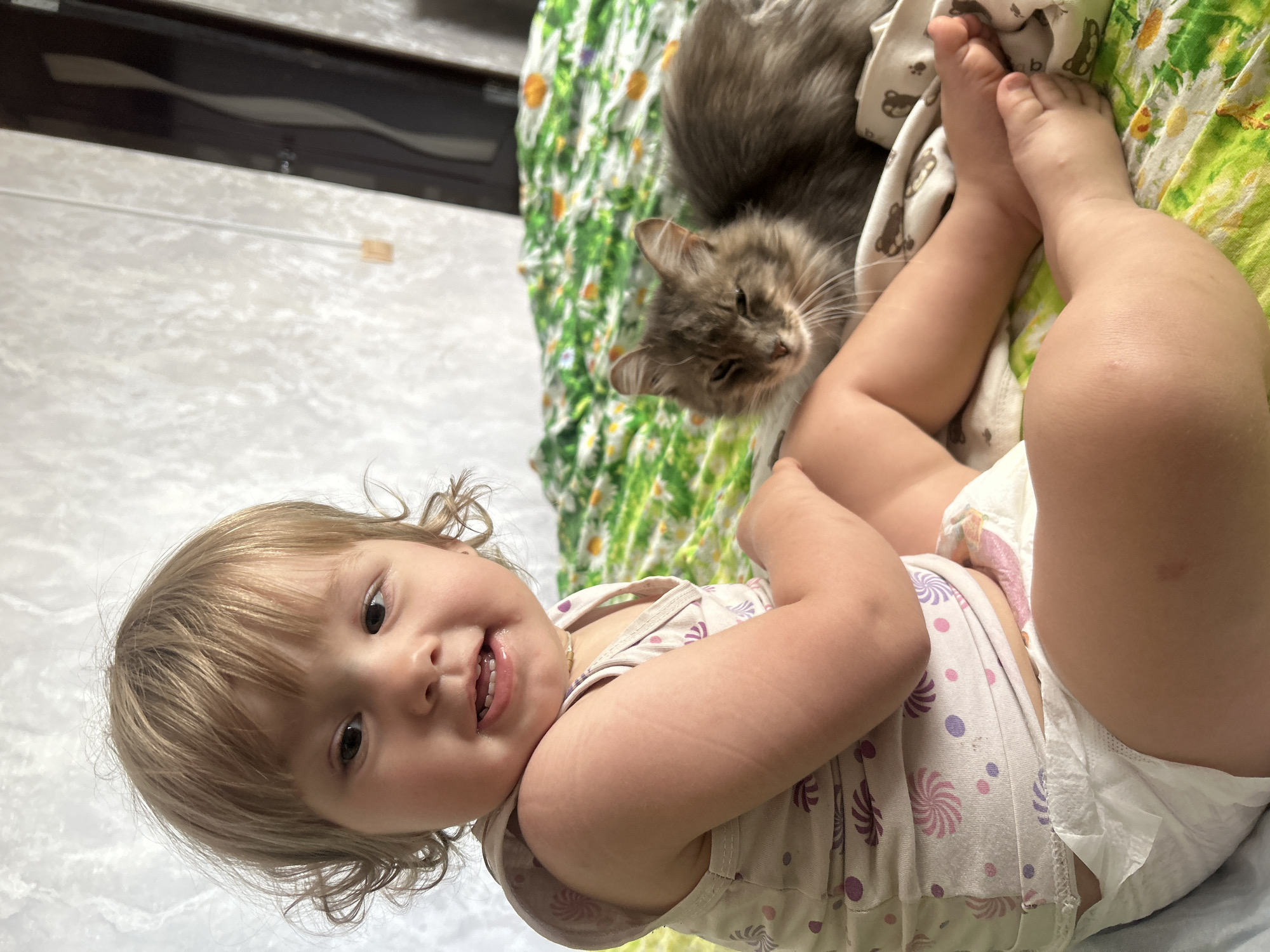 A young Caucasian child with blonde, curly hair and a light-colored tank top patterned with small, colorful circles and swirls sits on a bed with a floral-patterned green and white comforter. The child is facing forward and smiling, with their head tilted slightly. Their right arm is bent and resting on their lap, and their legs are extended. A fluffy, grey cat with green eyes is lying on the comforter to the right of the child, with its eyes partially closed. The background consists of a textured, off-white wall with a dark vertical object, possibly a door, to the right. The scene appears to be indoors, in a domestic setting. There is no indication of the location in the city of Olkhovka, Russia.