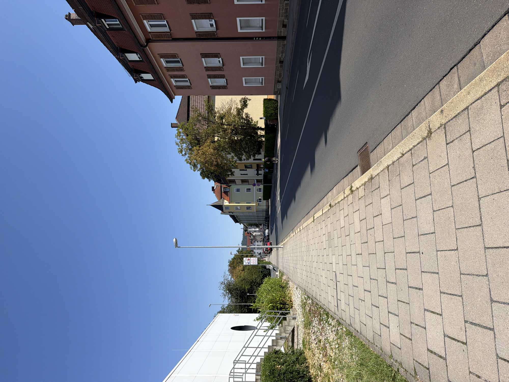 The image depicts a street scene in Bayreuth, Germany, on a clear, sunny day with a bright blue sky. The perspective is from the sidewalk, looking down a relatively wide, asphalt road.
On the left side of the image, a modern, white building with a large, dark circular opening dominates the foreground. Adjacent to this building, a concrete staircase with metal railings leads up to an entrance. The sidewalk in front of the building is paved with light-colored rectangular tiles. A strip of grass and gravel is visible between the sidewalk and the building.
The right side of the street is lined with multi-story buildings in various architectural styles, typical of a European city. The closest building is a reddish-pink structure with a mansard roof, multiple windows with wooden shutters, and a red-tiled roof. Further down the street, other buildings are visible, including a yellow building with a peaked roof and a green building with a tower-like element. Trees with autumn foliage are interspersed along the street.
A lamppost stands prominently on the sidewalk in the middle ground. There are a few cars parked or driving on the street, and two individuals are visible on bicycles further down the road, one wearing a yellow helmet. A street sign is also visible on the right side of the sidewalk. The road has white lane markings and a white arrow indicating a turn. A storm drain is present at the edge of the sidewalk. The overall impression is of a well-maintained urban environment during daylight hours.