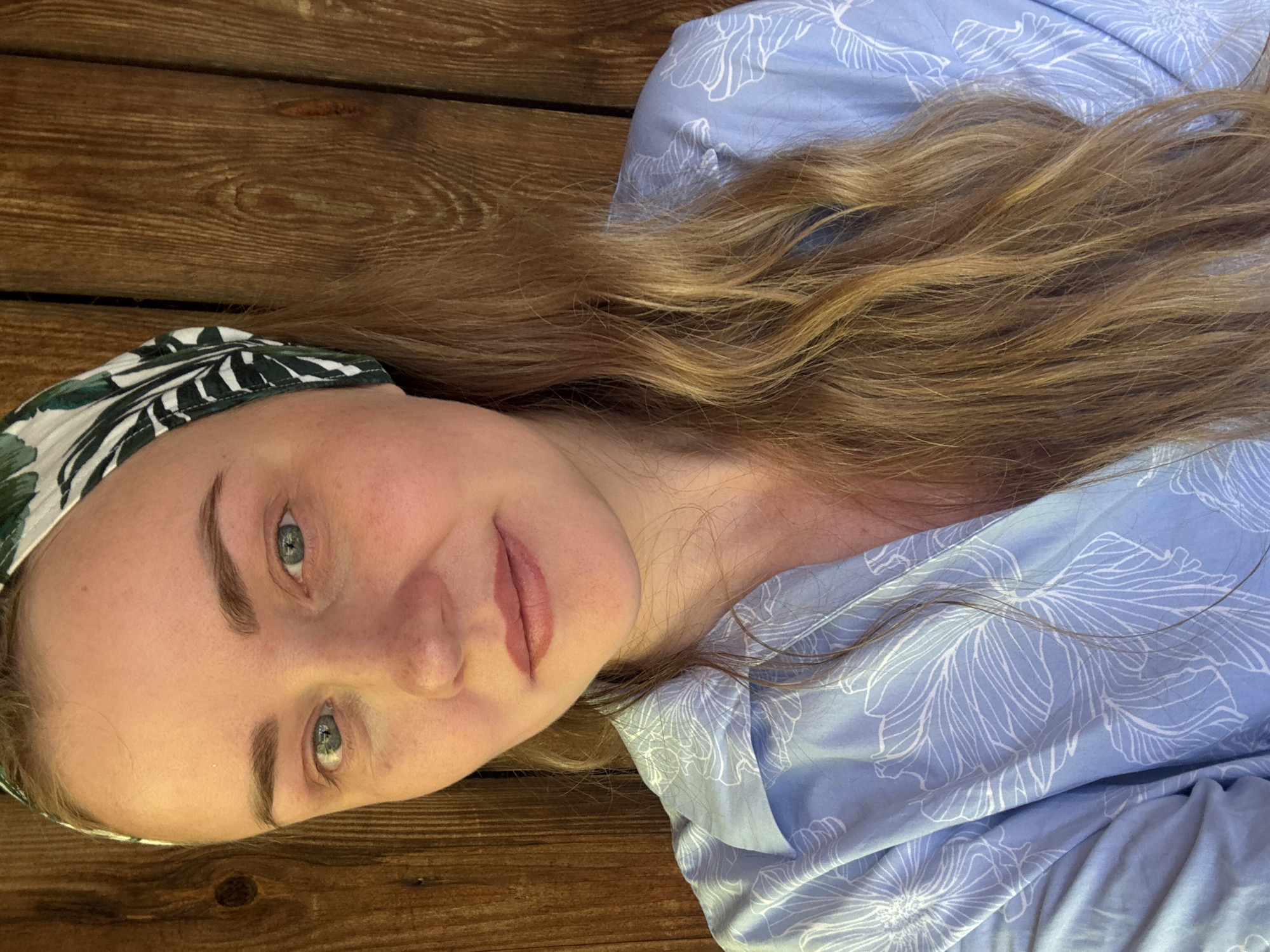 A fair-skinned woman with blue eyes and light brown hair is the primary subject. She is wearing a headband with a green and white leaf pattern and a light blue top with white floral designs. The woman is lying on a dark brown wooden surface. The environment suggests an indoor setting with the wood likely part of the floor or wall. The photo's location within the city of Chaikovsky, Russia, cannot be determined.
