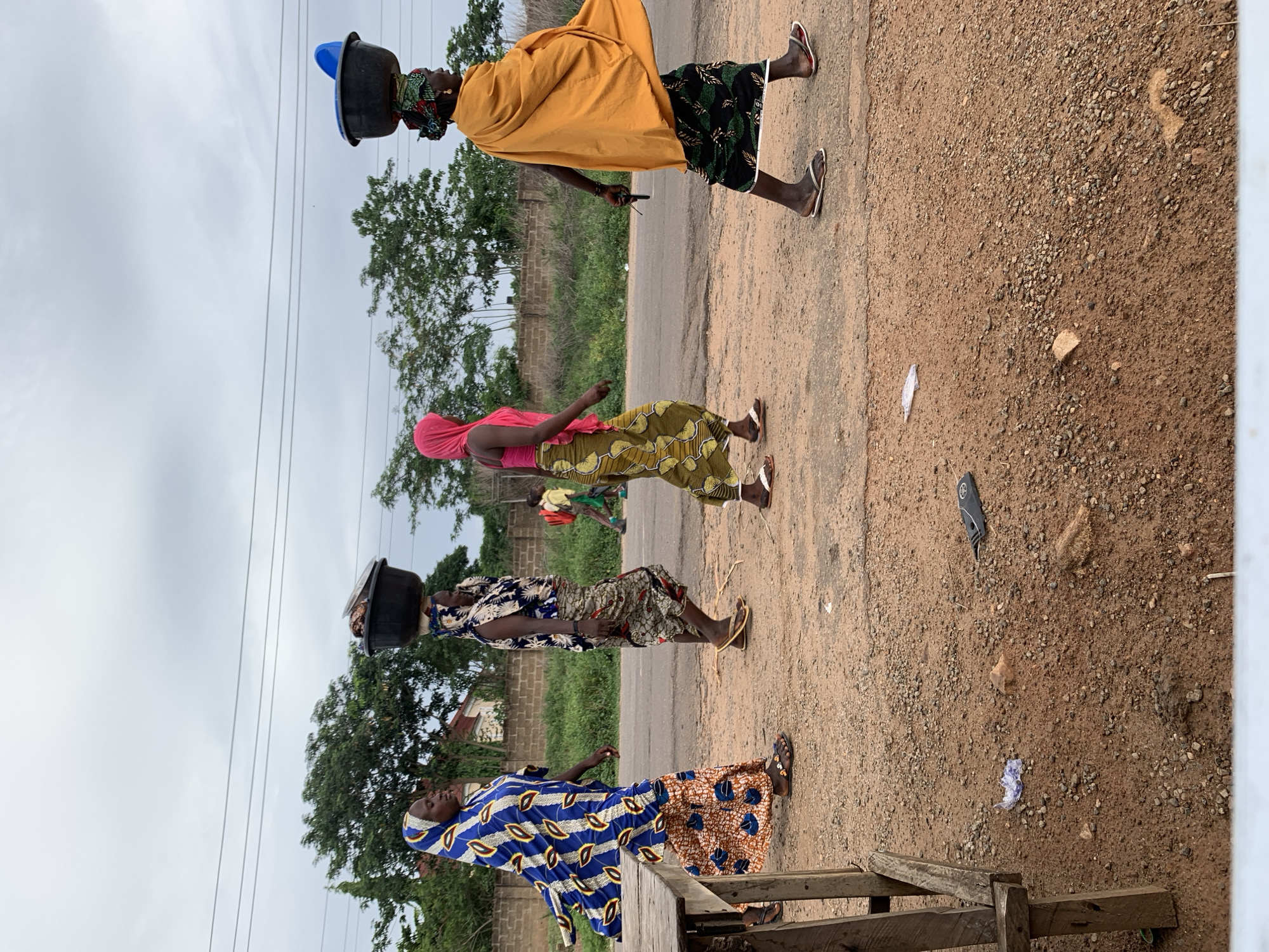In Gbeleko, Nigeria, four women are walking single file along a road. The woman at the front is wearing a yellow shawl, a green and black skirt, and sandals, and she has a black bucket on her head. The second woman is wearing a pink top, a yellow and green patterned skirt, and sandals, and she is carrying something green and red in her hands. The third woman has a black bucket on her head, and she is wearing a blue and white patterned top, a tan skirt, and sandals. The last woman is wearing a blue and brown patterned dress and sandals. The road is unpaved and there is a wooden structure in the foreground on the right side of the image. There are trees and bushes in the background, and power lines overhead.