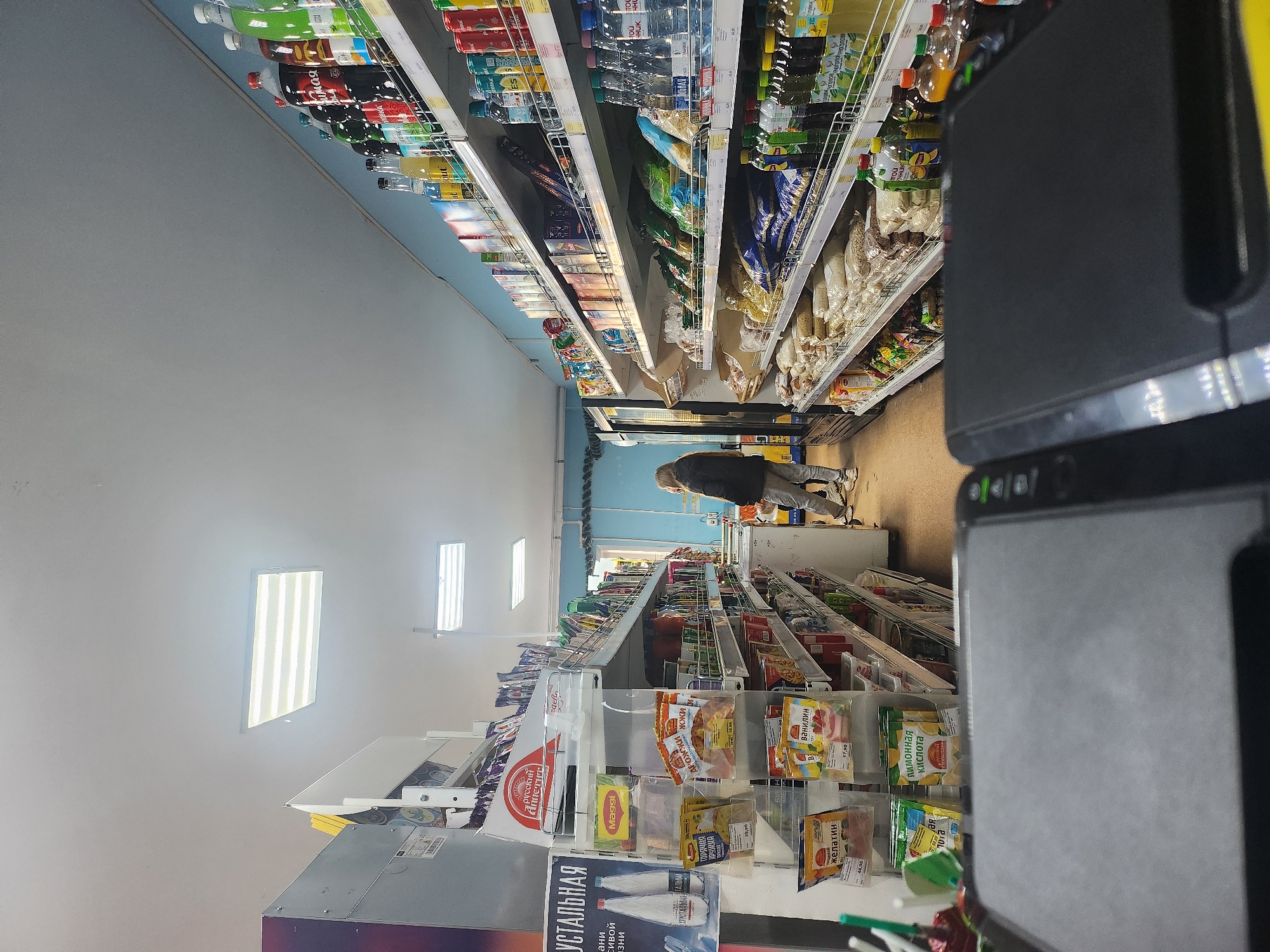 The original image is tilted 90 degrees clockwise, capturing an indoor scene within a convenience store or small supermarket in Krasnogorskoye, Russia.
The primary subject is a person in the middle ground, wearing a dark jacket and light-colored pants, bent over to view items on a low shelf. Their back is to the camera, and they appear to be shopping.
The store is lined with shelves fully stocked with various goods. On the left side of the frame, shelves display numerous bottles, likely beverages such as soft drinks and water, with visible brands like "HUK" and "ХРУСТАЛЬНАЯ" (Crystal). Further down these shelves are packaged goods, including what appears to be "русский Аппетит" (Russian Appetite) brand items.
On the right side of the frame, shelves also hold bottles of beverages, alongside bags of grains, cereals, and other packaged food items. In the closer foreground on this side, a display rack contains smaller packets of spices and baking ingredients, including "Maggi" products, "ГОРЯЧАЯ КРУЖКА" (Hot Mug) instant mixes, "ванилин" (vanillin), "желатин" (gelatin), and "лимонная кислота" (citric acid), with price tags like "39,90", "17,90", and "44,90" rubles visible. A large, dark piece of equipment, possibly part of a checkout counter or scanner, dominates the far right foreground, partially obstructing the view of the shelves beyond.
The store is lit by several bright rectangular fluorescent light panels mounted on the ceiling or upper wall, suggesting an indoor setting with artificial illumination. The walls are a plain light grey or off-white, and the floor is a darker tone. The atmosphere appears to be that of a typical, functional grocery retail environment.