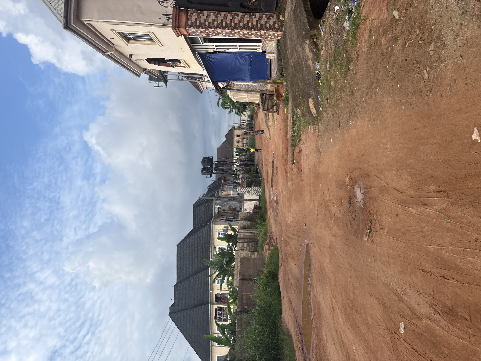The image depicts an unpaved, reddish-brown dirt road in Obosi, Nigeria, with evidence of recent rainfall in the form of puddles and muddy patches. On the right side of the road, a two-story building with a light-colored facade and a grey, tiled roof is visible. This building features a ground-floor storefront covered by a blue tarp. Adjacent to it is a brick pillar with a decorative pattern. Further down the road, several other residential buildings with grey tiled roofs are visible.
On the left side of the road, a long, low-lying building with a dark grey, tiled roof is prominent, flanked by mature green plants, including banana trees. A low brick wall separates this building from the road. In the distance, two individuals are walking along the road, one wearing a yellow shirt and the other a blue garment. Overhead, a cloudy sky with patches of blue dominates the upper portion of the image. There are power lines visible in the upper left quadrant. Scattered debris and litter are present along the sides of the road.