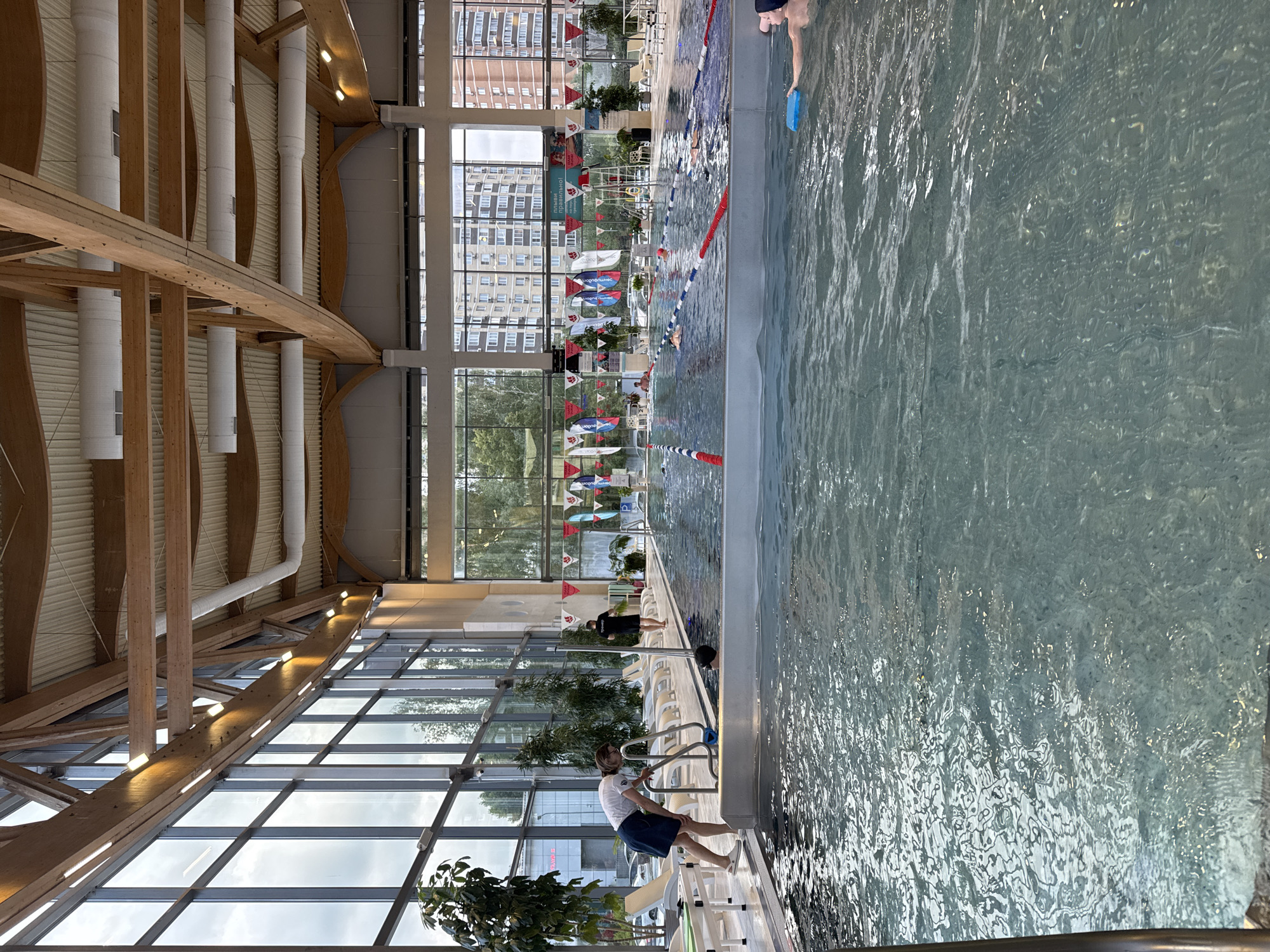This is an indoor swimming pool facility with a large swimming pool. The pool is divided into lanes by ropes and buoys. Several people are visible in the water, some are swimming, and one person is holding a blue kickboard. A woman in a white shirt and blue skirt is standing at the edge of the pool, near a ladder. Another person wearing a black shirt with "ТРЕНЕР" (trainer) written on the back is walking near the pool. The pool area is enclosed by large windows, allowing natural light to enter. The architecture features exposed wooden beams in the ceiling and modern lighting. Outside the windows, residential buildings and trees are visible, suggesting an urban or suburban setting in the city of Sovkhoza imeni Lenina, Russia. The water in the pool is clear, and there are lounge chairs and plants around the perimeter.