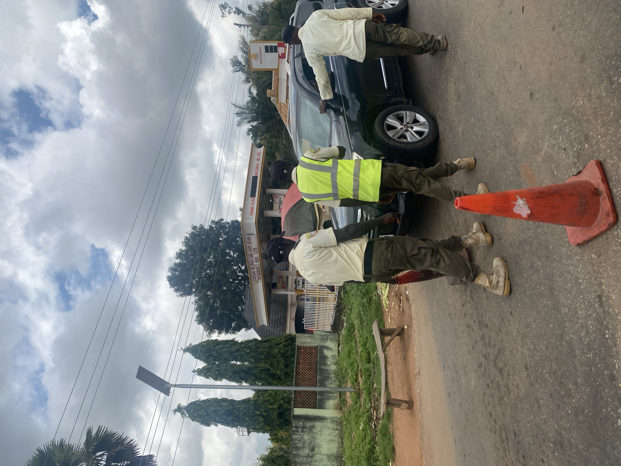 The image depicts a roadside scene in Tataura, Nigeria, likely near a gas station. Three men, appearing to be officials, are present. One man in a light-colored uniform with a dark cap stands in the foreground, facing away from the camera. Another man, wearing a high-visibility yellow vest over a similar uniform and a dark cap, stands next to a dark-colored SUV, seemingly interacting with it. A third man in a light-colored uniform and dark cap stands to the right of the SUV, with his hand on the vehicle's door. An orange traffic cone is visible on the asphalt road in the foreground. In the background, a gas station with signage is visible, along with trees and power lines. The sky is cloudy.