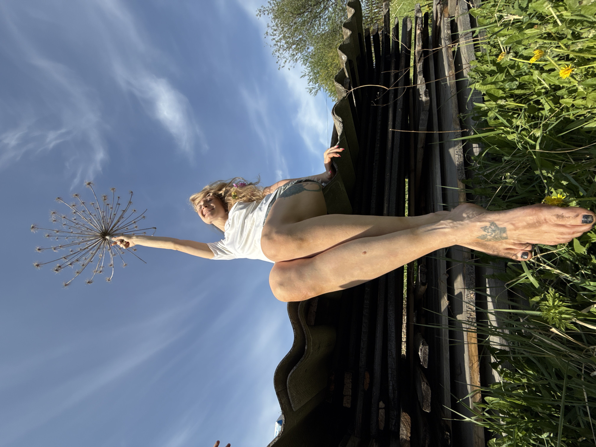 The image depicts a woman lying on what appears to be corrugated roofing material stacked vertically. She is wearing a white t-shirt and shorts, with a flower in her hair. She holds a large dried seed head in her outstretched arm against a blue sky with wispy clouds. The setting includes grass and dandelions on one side and a tree line in the background. The scene is located in Nizhnyaya Chermoda, Russia.
