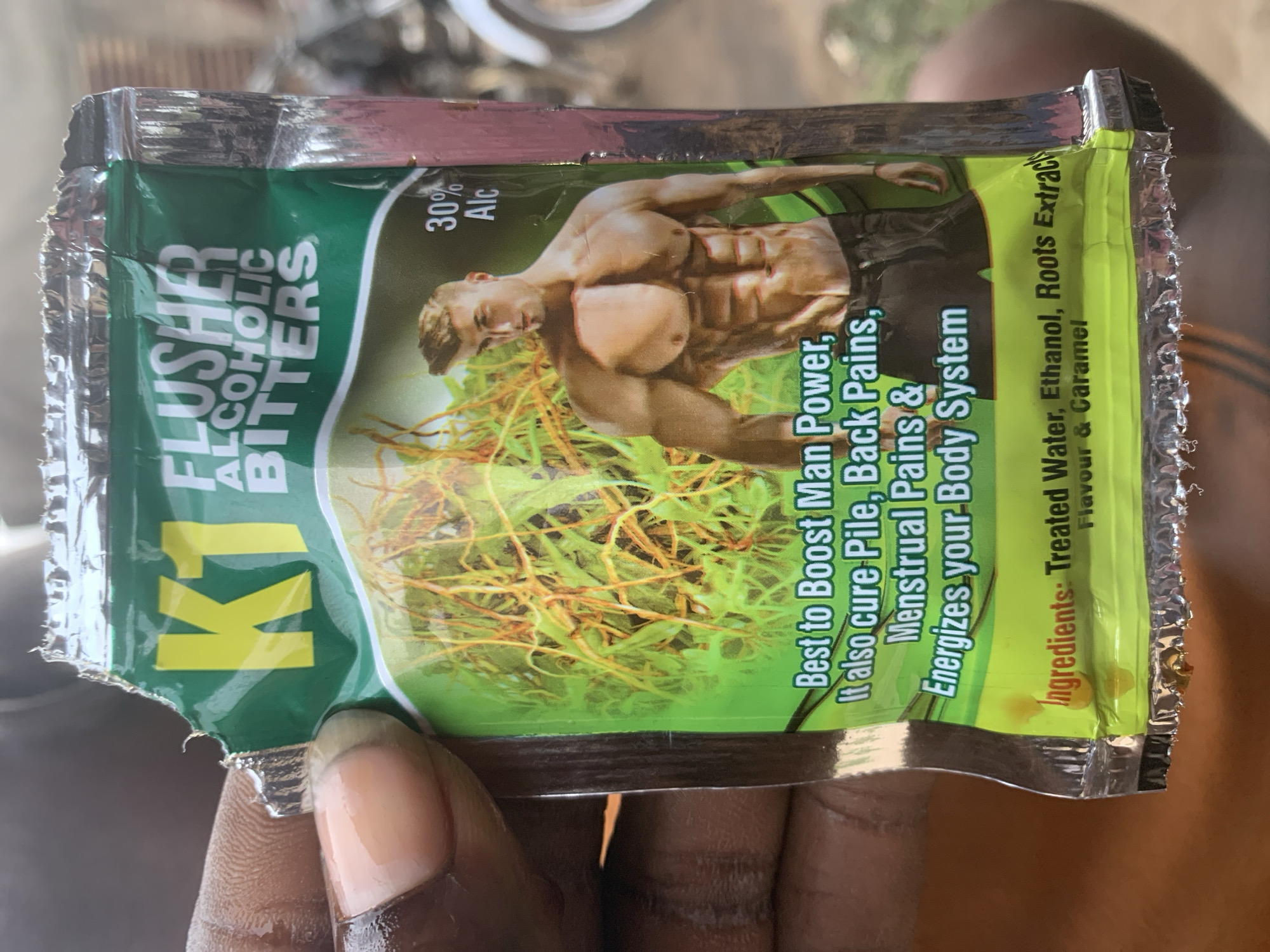 In Shagamu, Nigeria, a person's hand holds a sachet of "K1 FLUSHER ALCOHOLIC BITTERS." The sachet is primarily green with yellow lettering, featuring an image of a muscular man. Text on the sachet indicates it is "Best to Boost Man Power, It also cure Pile, Back Pains, Menstrual Pains & Energizes your Body System." The ingredients are listed as "Treated Water, Ethanol, Roots Extracts, Flavour & Caramel" with 30% Alcohol. The sachet appears to be torn open at the top and bottom edges. The background is out of focus, but suggests an outdoor setting with some vehicular elements visible.