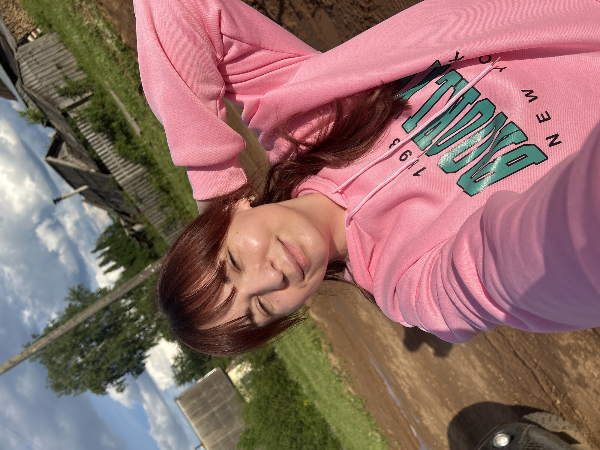 The image is a selfie of a woman lying outdoors. She has reddish-brown hair with bangs and is wearing a pink hoodie with green lettering. The background shows a rural environment, with what appears to be a dirt or muddy path, grass, and old wooden buildings reminiscent of a village. The sky with clouds is visible above. Based on the information provided, the location is Фоки, Russia.