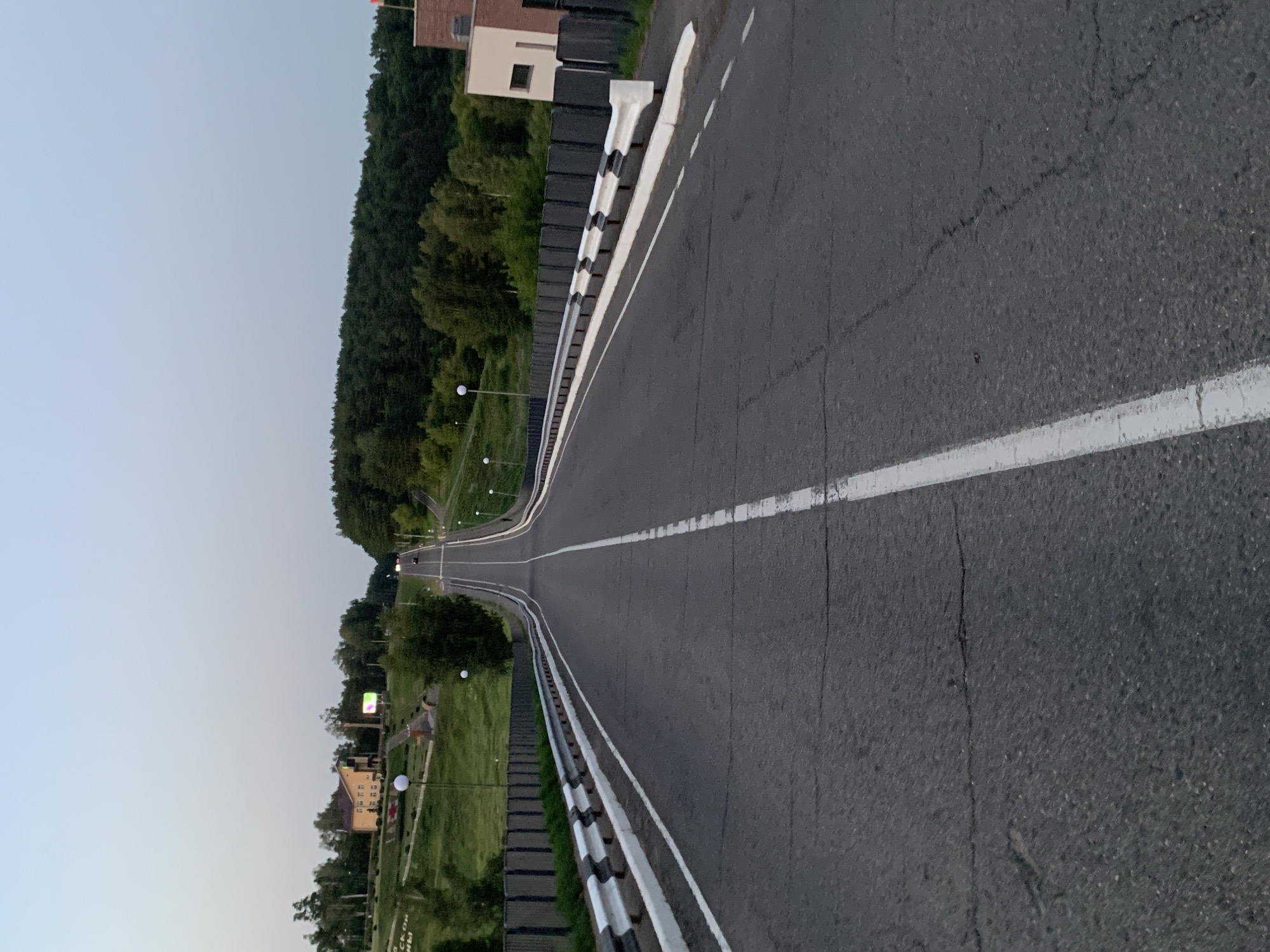 The image shows an asphalt road leading uphill, with a clear white line marking the center. Guardrails with black and white striped reflective elements line both sides of the road. In the distance, the road curves and disappears over the crest of the hill. The sky is a pale blue, suggesting it is either early morning or late evening.
On the left side of the road, a grassy slope rises, with a yellow building and some trees visible. There are also lampposts with white spherical lights on the slope. In the far distance, a dense forest covers the landscape. On the right side, there is a modern-style building with a beige facade and dark windows.
The road appears to be in a somewhat rural or suburban setting, possibly on the outskirts of a city or town. The presence of the guardrails and the clear lane markings suggest it is a well-maintained public road. The overall impression is one of quiet solitude, with no people or vehicles immediately visible in the foreground. Based on the visual cues, this road is located in the city of Kapyĺ, Belarus.