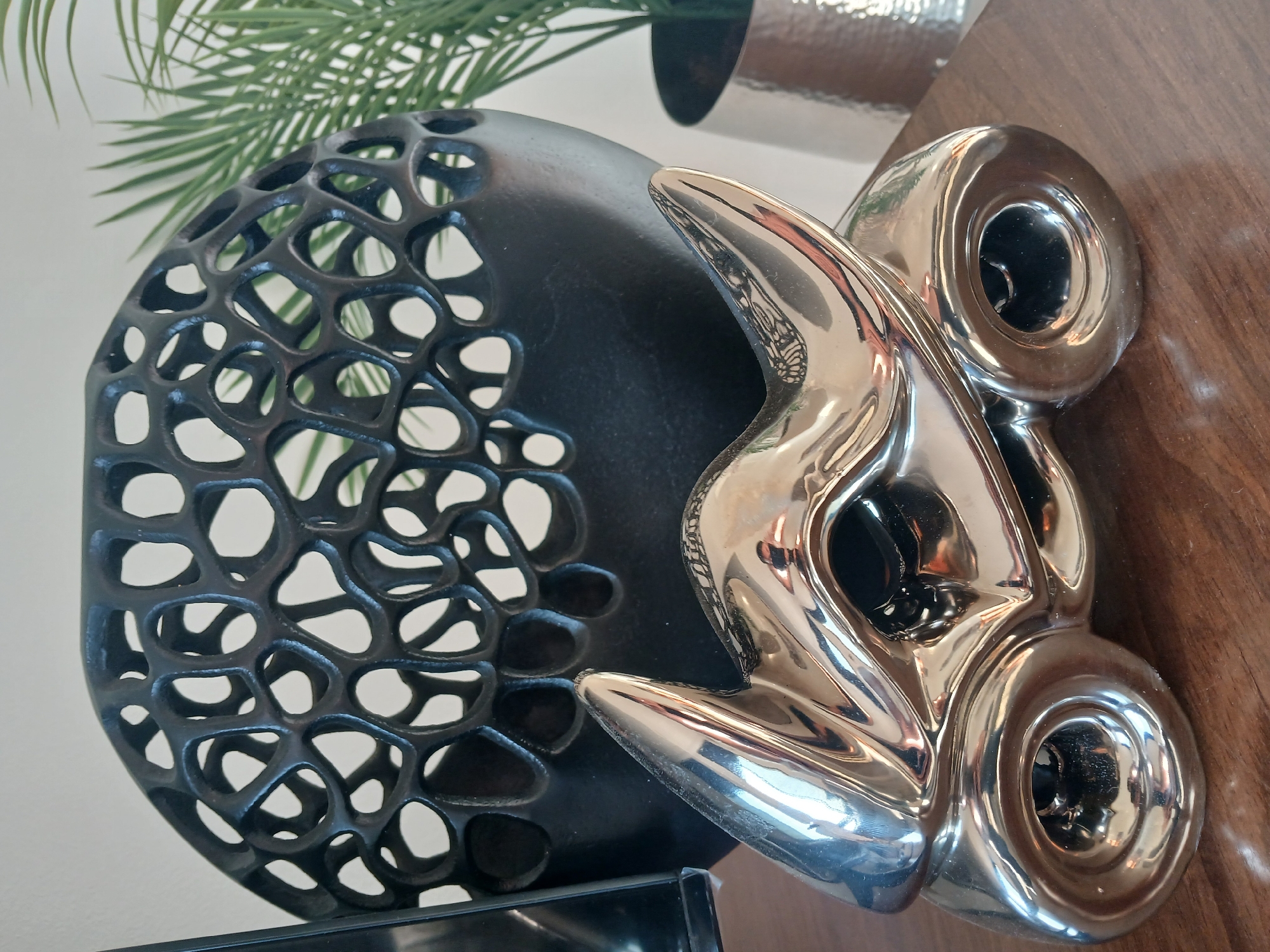 The image presents a still life arrangement of decorative objects on a wooden surface. A black, spheroid vase with an irregular, lattice-like pattern of cutouts is positioned to the left. Beside it is a silver-colored abstract sculpture with two rounded elements featuring circular openings. Behind the vase, there's a glimpse of a green plant, presumably an artificial decorative piece. The background is a light-colored wall. The location and specific details of the city are unknown.