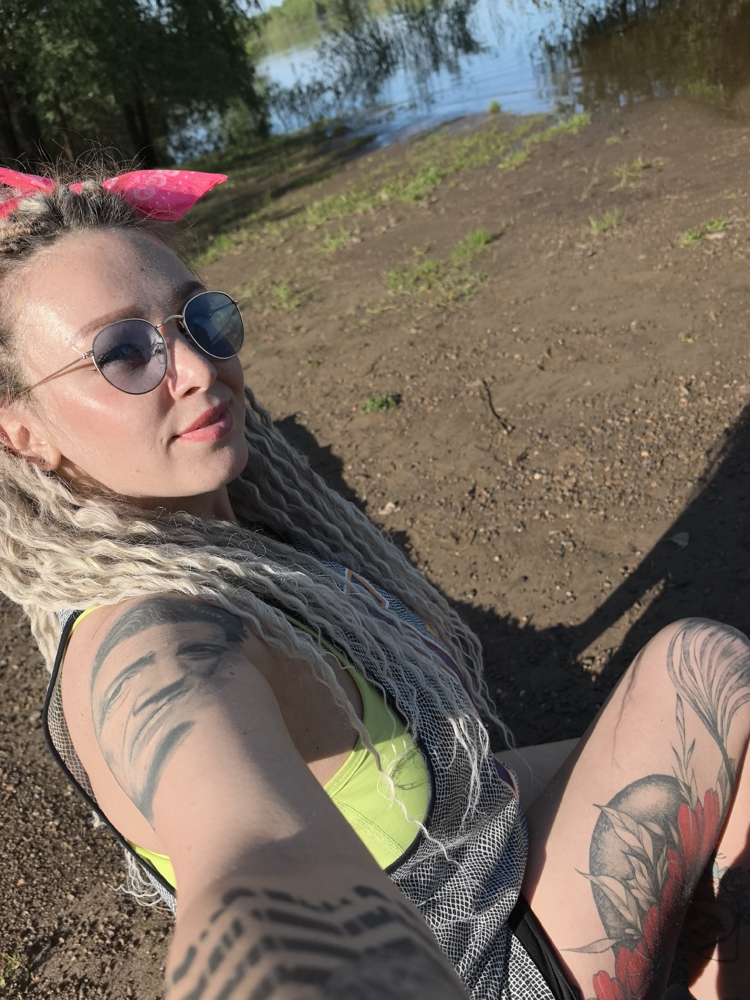 A woman with long blonde dreadlocks and sunglasses is sitting on the ground near a body of water. She has a pink bow in her hair and tattoos on her arms and legs. The location is Noviy, Russia. The ground is dirt and gravel. The water is in the background, with trees along the shoreline.