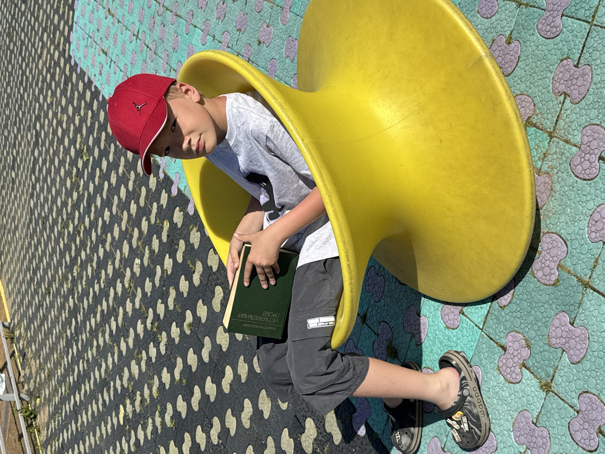 A child sits inside a yellow playground structure, reading a book. The child wears a red baseball cap, a gray t-shirt, gray shorts, and Crocs-style shoes. The book has Cyrillic text on its cover. The playground surface is patterned with interconnected designs in shades of teal, purple, and gray. Another surface with a different pattern, black with light shapes, lies nearby. The scene is in Синьково, Russia.