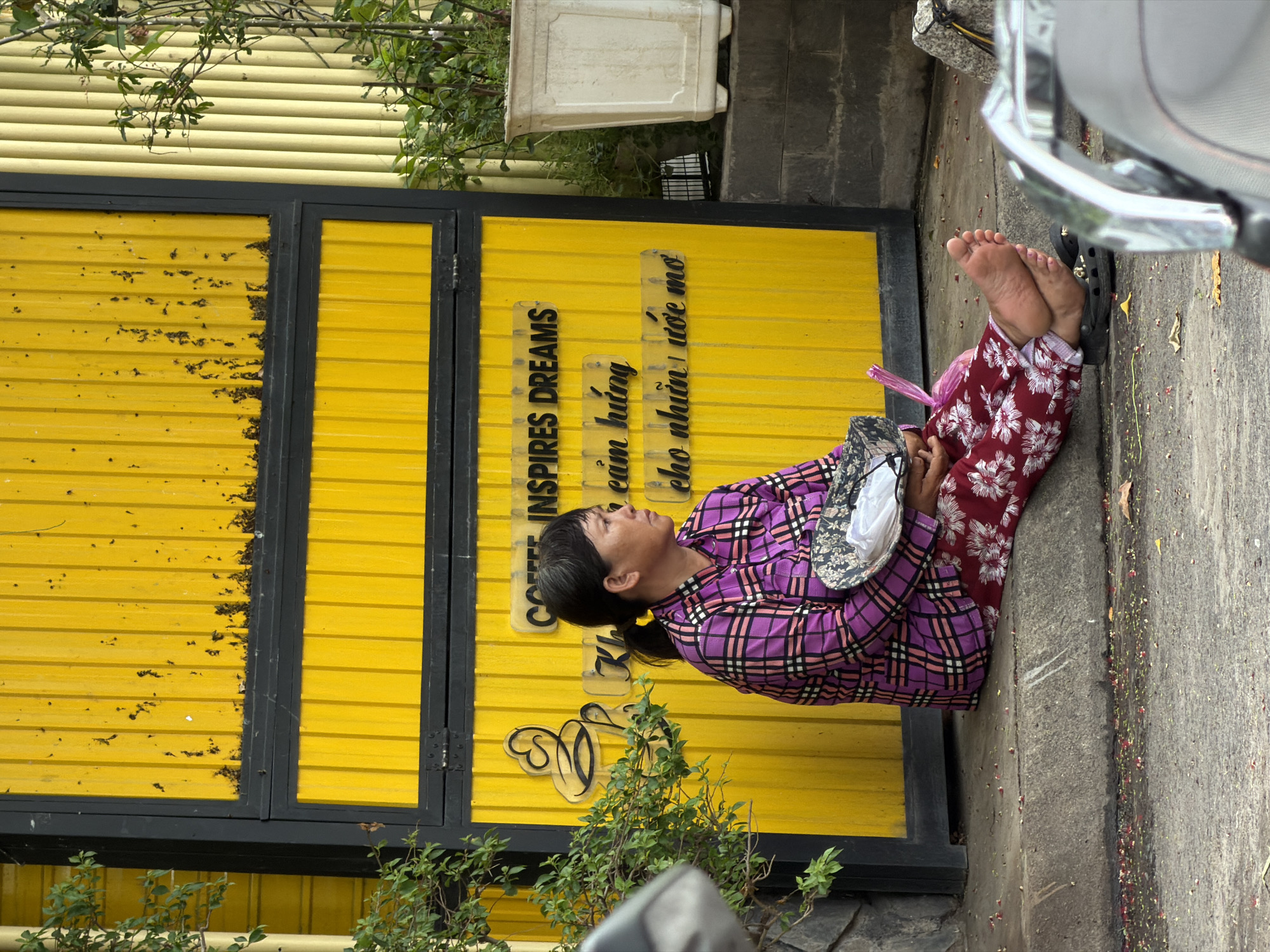 An outdoor, eye-level shot captures an individual resting on a concrete sidewalk next to a vibrant yellow corrugated metal wall or door in Nha Trang Ward, Vietnam. The person, likely female, is seated on the ground with her legs extended, wearing a purple and black plaid long-sleeved shirt and red pants with white floral patterns. She is barefoot, with a single black sandal positioned near her feet. She holds a patterned object, possibly a hat, which contains a white face mask, and a pink plastic bag lies beside her. Her expression is calm or contemplative.
The bright yellow wall features black framing and shows signs of wear, including dark smudges. Mounted vertically on the wall are three clear acrylic signs. The first reads "INSPIRES DREAMS" in English. The subsequent two signs are in Vietnamese: "cảm hứng" (meaning "inspiration") and "cho những ước mơ" (meaning "for dreams"), together conveying a message related to inspiring dreams. A decorative cursive symbol is also visible below the signs.
The setting is an urban street or alleyway under bright daylight. Green foliage from plants or trees is visible at the top of the wall and near the ground level. A white rectangular planter is seen in the upper background. The concrete ground where the person sits is marked with small, colorful specks, possibly dried flowers or confetti. To the right of the person, the chrome bumper and part of a white vehicle, likely a motorcycle or scooter, are partially visible.
