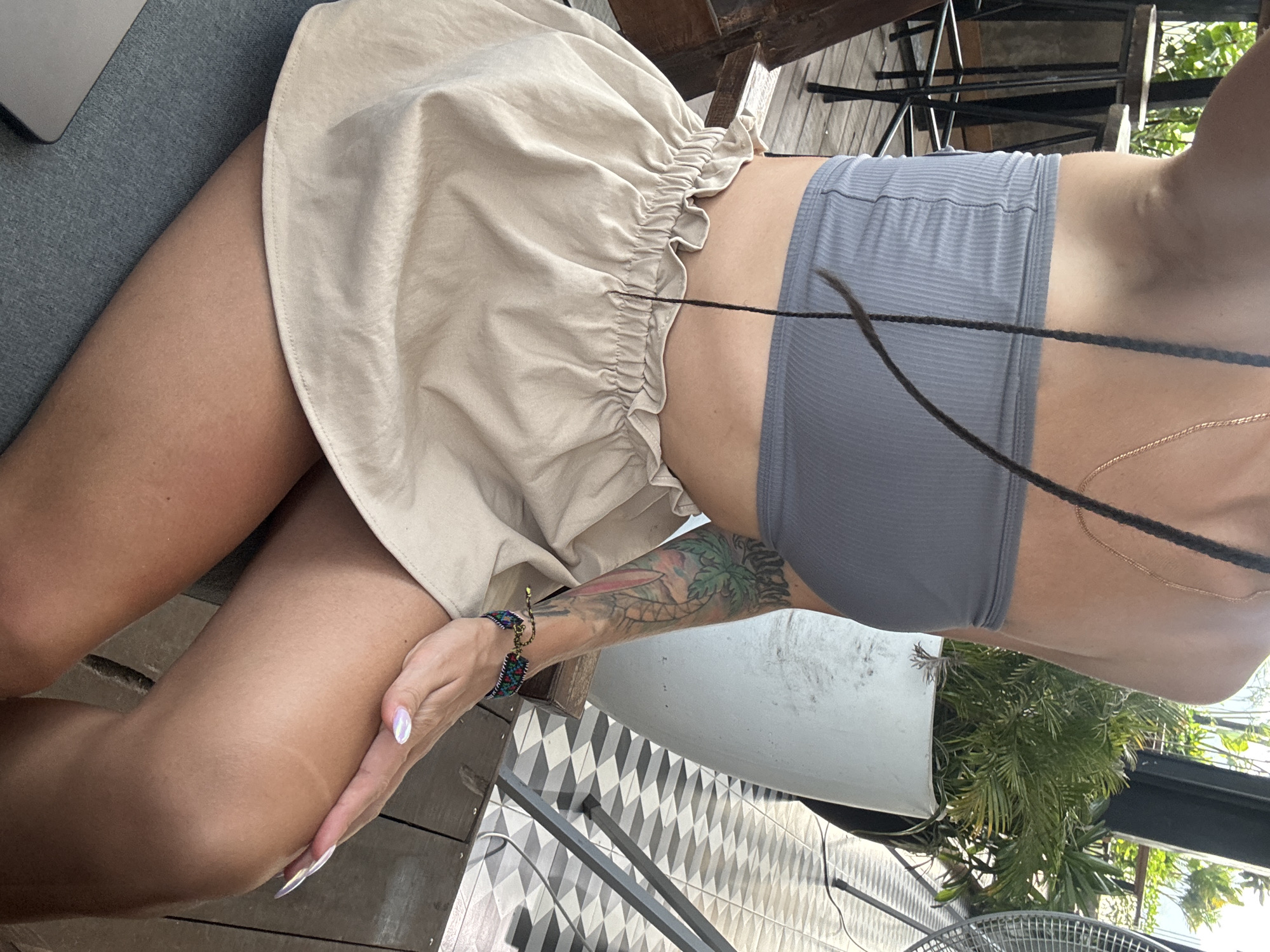 A person is depicted lounging in what appears to be an outdoor or semi-outdoor setting. The image primarily focuses on their mid-section, legs, and left arm. They are wearing a light beige, ruffled mini-skirt or skort paired with a form-fitting gray ribbed crop top or bralette. A gold chain necklace is visible around their neck, and multiple long, dark braided hair strands cross their torso.
On their left forearm, a prominent, colorful tattoo features tropical elements, including palm trees and what might be a surfboard, set against a colorful sky. A vibrant, multi-colored beaded bracelet adorns their left wrist, and their fingernails are long with a pearlescent polish. One hand rests casually on their thigh.
The setting includes a gray cushioned surface, possibly a sofa or bench, where a silver laptop is partially visible. The background reveals a mix of natural and architectural elements: wooden planks, a white plant pot filled with green leafy plants, black metal railings or furniture, and a distinctive black and white geometric tiled floor. Further back, more green foliage and structural elements of a building are visible. A fan is partially seen at the bottom right. The scene is brightly lit, suggesting it is daytime. No text is visible in the image. The context for the location is Hà Giang 1 Ward, Vietnam.
