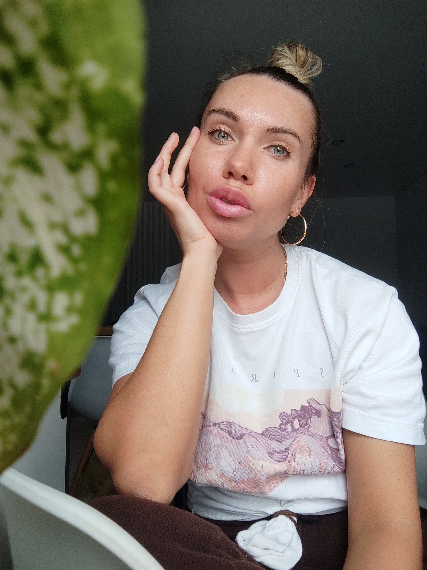 A young woman is posing for a selfie indoors. She has fair skin, light brown hair styled in a top knot, and is wearing gold hoop earrings. She is wearing a white t-shirt with a graphic print. Her right hand is supporting her chin, and she is pouting. A large, green and white patterned leaf is positioned on the left side of the frame, partially obscuring the view. The background is dimly lit. There is no indication of a specific location in Votkinsk, Russia. The scene is simply a casual indoor shot.