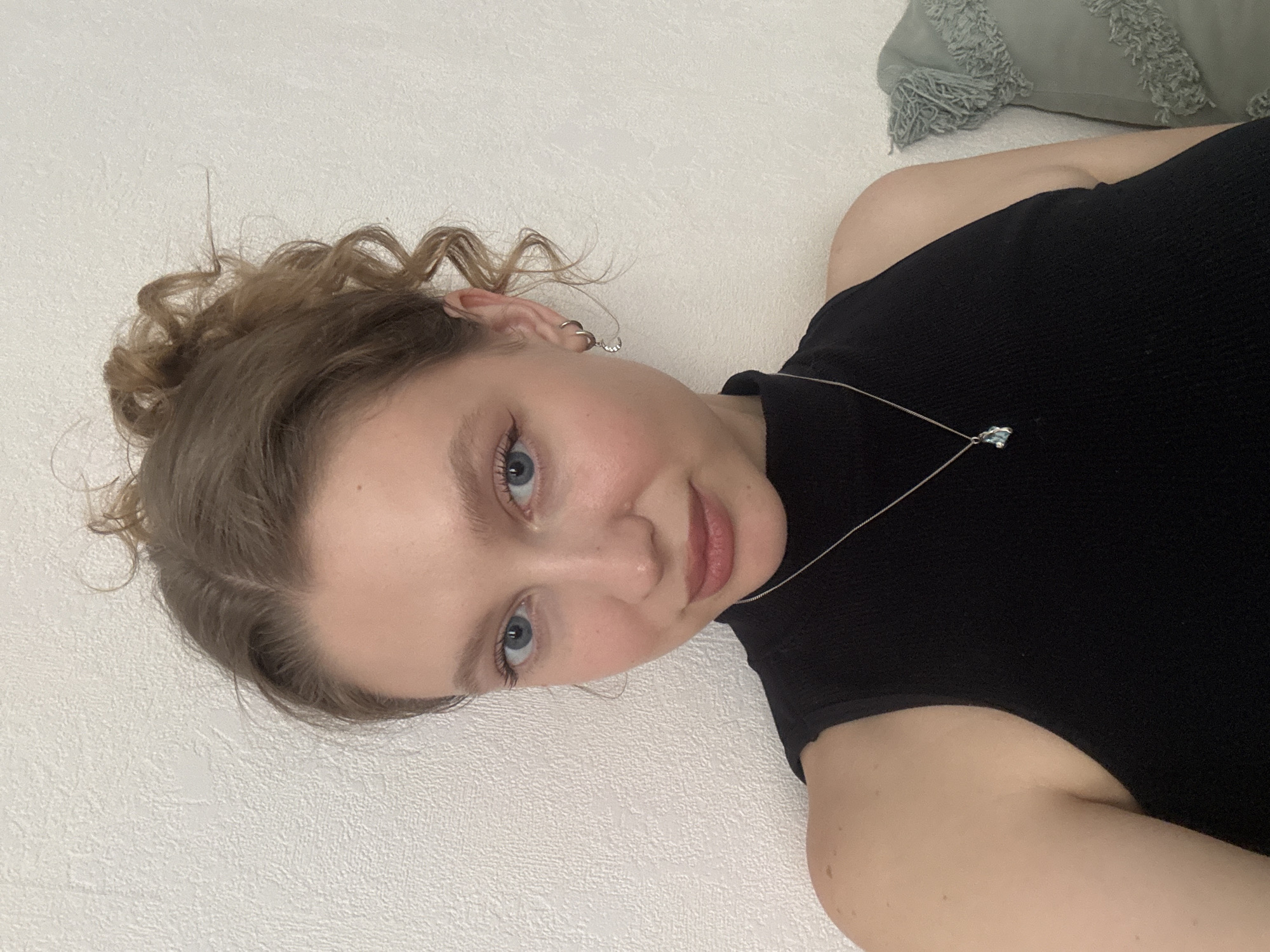 A young woman with light skin, blue eyes, and light brown hair styled in a messy bun is looking at the camera. She is wearing a black sleeveless top and a silver necklace with a light blue pendant. She has on eyeliner and light makeup. She is wearing small silver hoop earrings, one with a moon charm. The background is a textured white wall. To the right of the woman, a light green pillow with fringe is visible. The image appears to have been taken indoors. There is no information to determine the location in the city of Izhevsk, Russia.