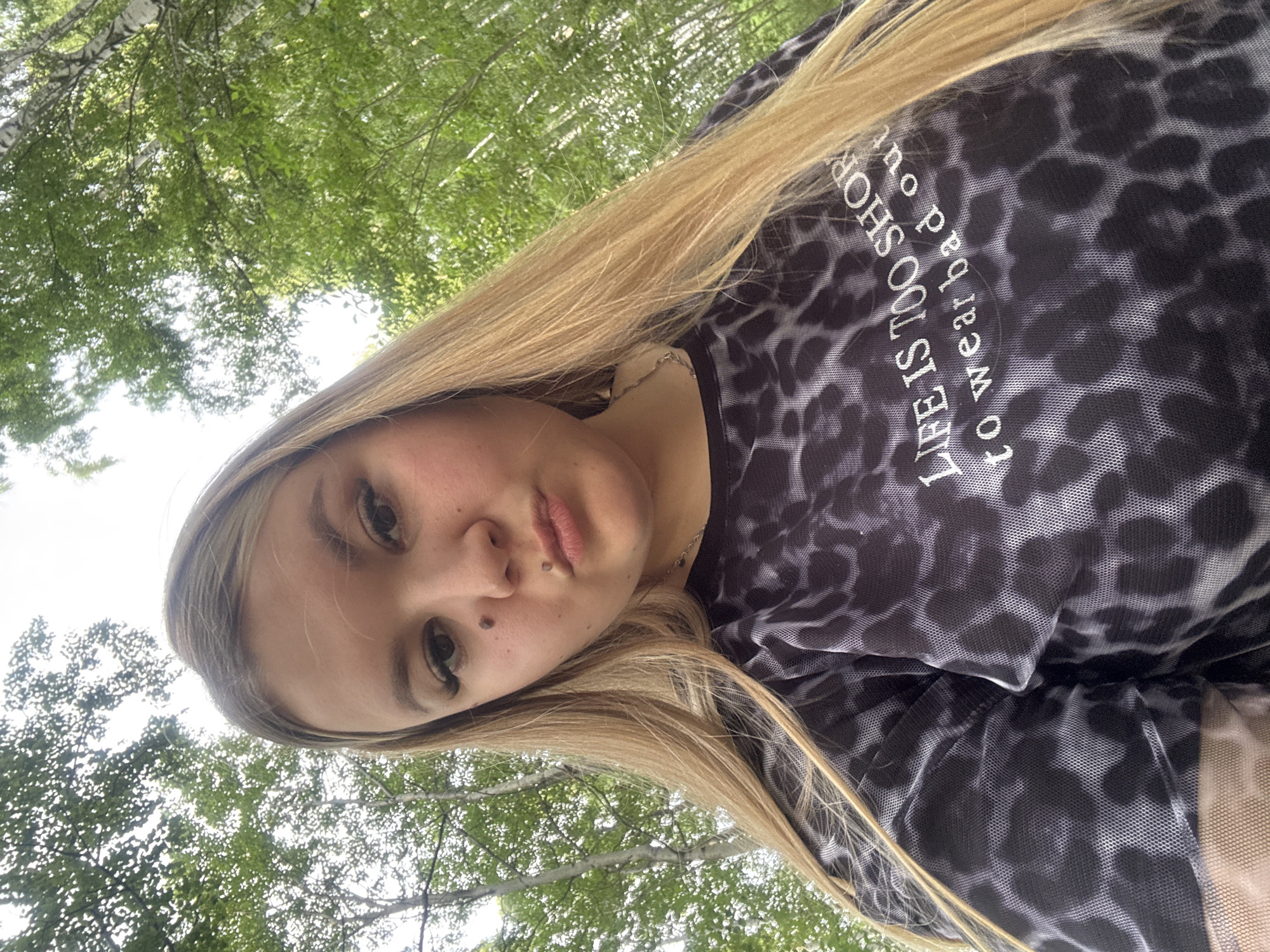 A young woman with long blonde hair is taking a selfie outdoors. She is looking directly at the camera with a neutral expression. She is wearing a black and grey leopard print top. The background consists of green trees and a bright sky, suggesting it is daytime and the weather is pleasant. The image is taken from a low angle, looking up at the woman. There is text on her top that reads "LIFE IS TOO SHORT TO WEAR BAD OUTFITS". The location context is Chaikovsky, Russia.