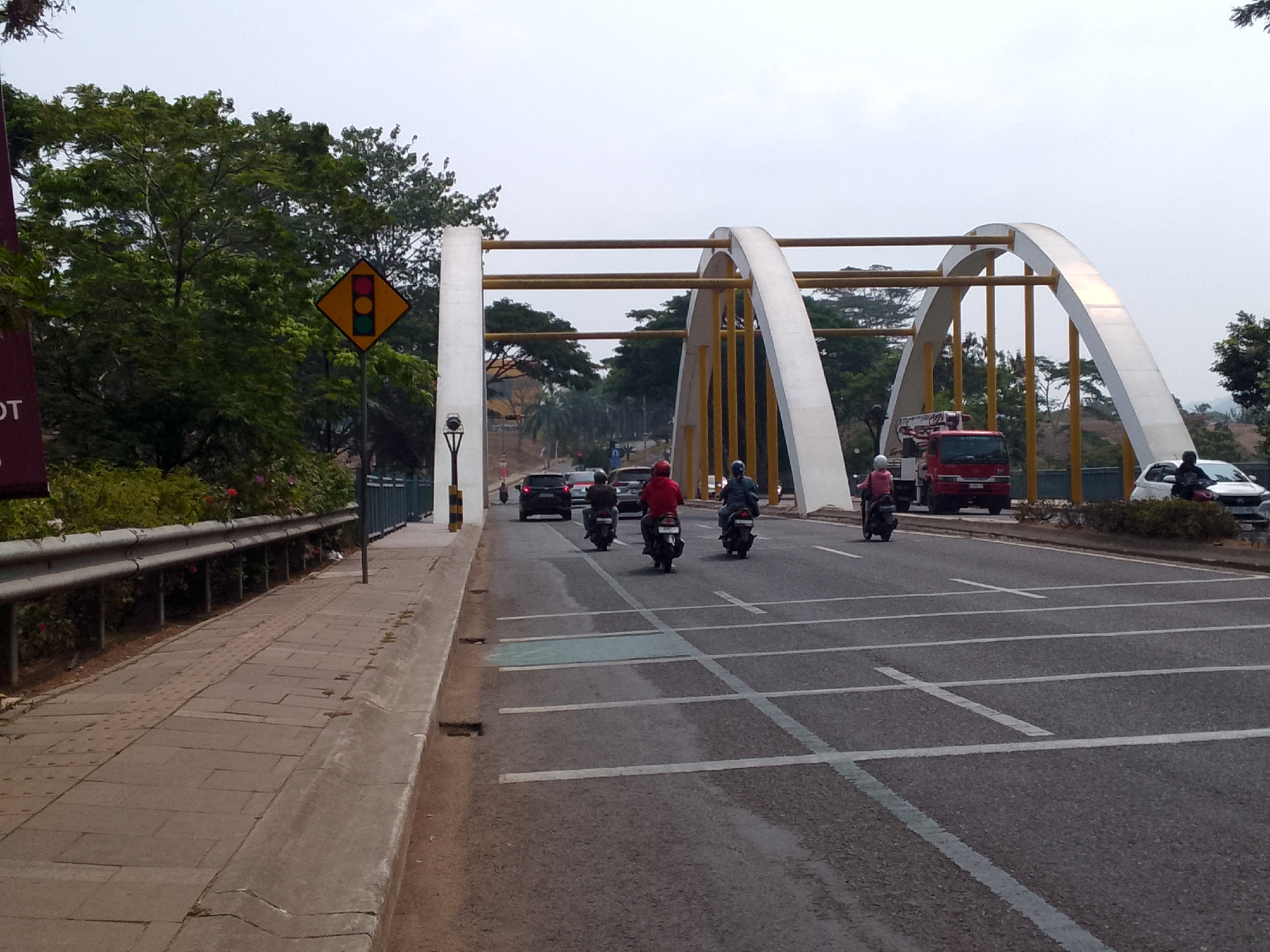 In Padalarang, Indonesia, on a cloudy day, a traffic scene unfolds beneath a distinctive arched bridge with yellow and white supports. On the right side of the road, several motorcycles and their riders are captured in motion. One motorcycle with a rider in a red jacket is positioned in the foreground, facing away from the camera. Another motorcycle, with a rider in blue, is situated slightly ahead and to the left of the first. A third motorcycle, with a rider in a pink jacket, is seen further to the left, moving in the same direction. To the right of these motorcycles, a white car is parked. In the background, a red fire truck is visible on the road. The road itself is marked with white lines, indicating lanes and crosswalks. On the left side of the image, a sidewalk runs alongside the road, bordered by a metal railing and lush green vegetation. A traffic signal sign, displaying red, yellow, and green lights, is prominently placed on the left side of the road, near the beginning of the sidewalk. The overall environment suggests a typical urban setting with moderate traffic and infrastructure.