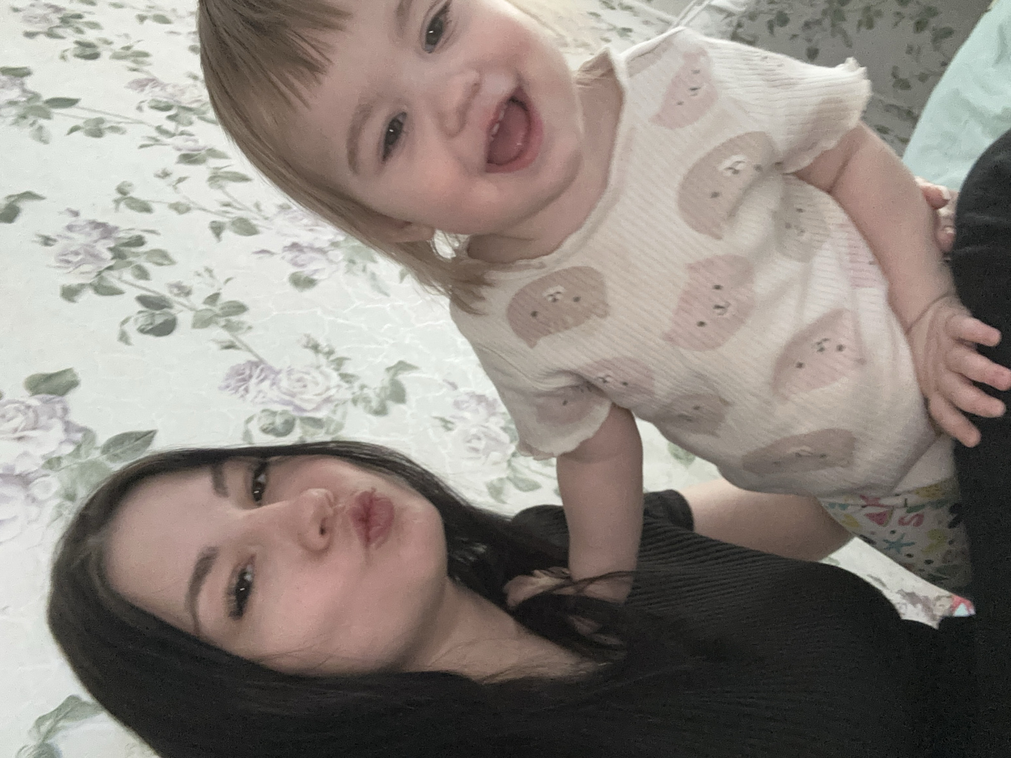The image displays an indoor scene in Chaikovsky, Russia. Two individuals are positioned against a light-colored wall adorned with wallpaper featuring a pattern of light purple roses and green leaves. On the left, an adult female with long, dark hair, wearing a black ribbed top, looks towards the viewer with pursed lips. Her right arm is positioned next to the child. On the right, a young child with light blonde hair and bangs, wearing a light-colored, ribbed long-sleeved top with a pattern of pink and grey animal faces, smiles broadly at the viewer with an open mouth. Partially visible beneath the child are colorful patterned pants and a light blue fabric, possibly a blanket or bedding.