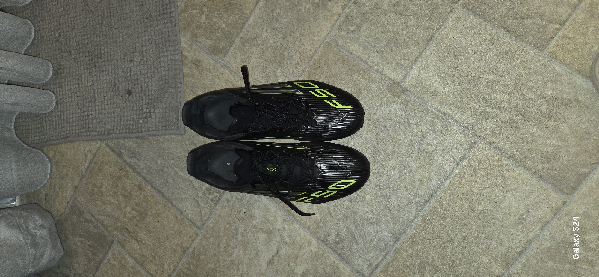 The image displays two black athletic shoes, likely soccer cleats, positioned on a light brown tiled floor. The shoes are oriented lengthwise, with one shoe pointing towards the top of the frame and the other pointing towards the bottom, creating a symmetrical arrangement. Both shoes feature prominent bright yellow-green "F50" lettering on their sides and matching stripe patterns.
To the left of the shoes, a grey, textured, woven floor mat is visible. Further to the far left, a light grey, possibly upholstered, object is partially seen, suggesting furniture or another soft furnishing. The tiled floor has visible grout lines and exhibits a subtle mottled texture.
No activity is taking place; the shoes are stationary. The scene appears to be an indoor setting, likely a home, based on the flooring and mat. The lighting is uniform, not indicating a specific time of day. The shoes appear to be in good condition.
Visible text includes "F50" on the side of both shoes. A "Galaxy S24" watermark is present in the bottom right corner of the image, indicating the device used for capture. The provided location context is Laval, Canada, though no specific visual cues in the image confirm this.