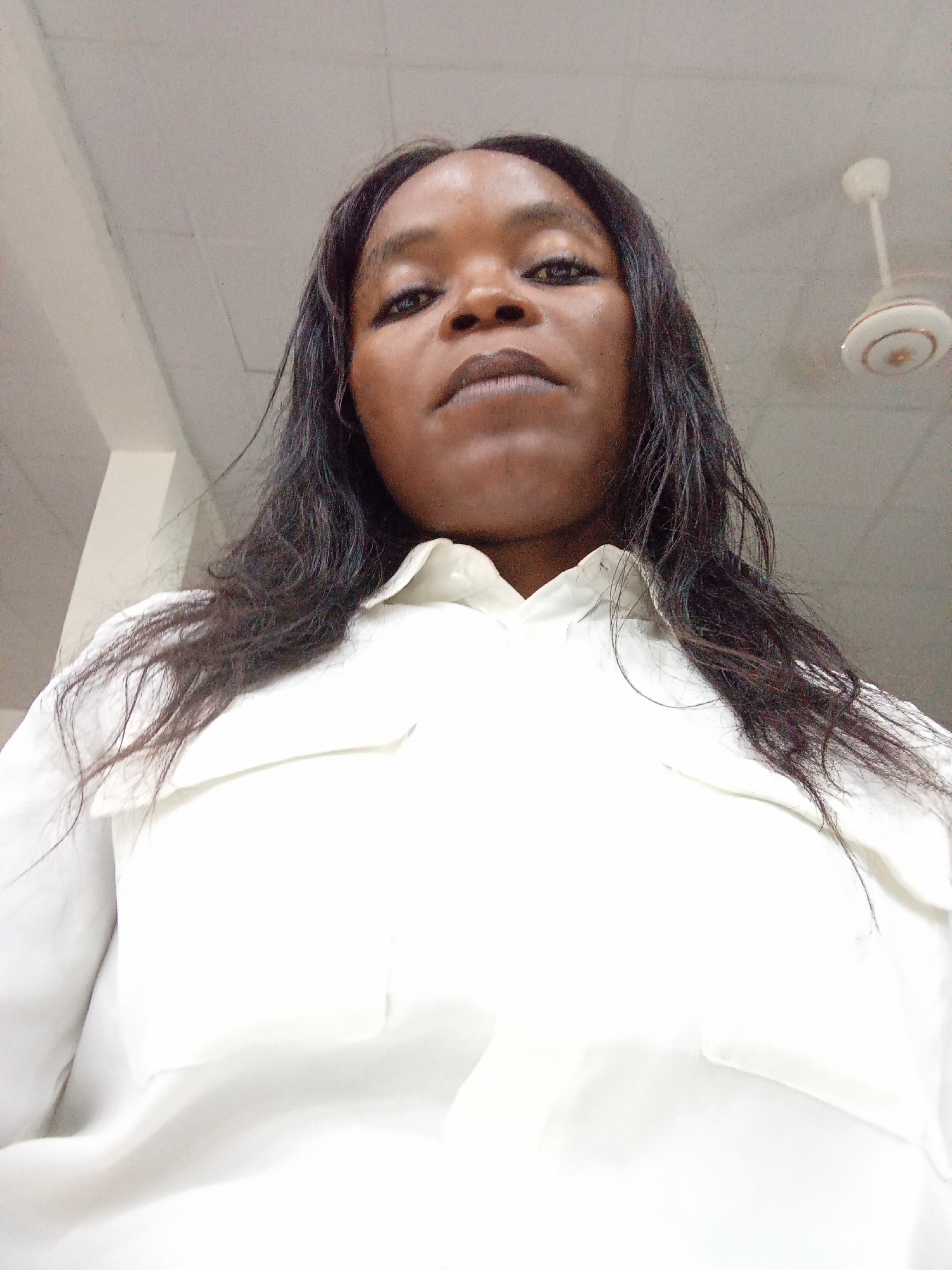 A waist-up, low-angle, outdoor selfie shows a Black woman with long, dark, straight hair. She is wearing a white collared button-up shirt. Her head is tilted slightly back and her gaze is directed towards the camera. The background appears to be an indoor ceiling with lighting fixtures and a ceiling fan visible. The scene is likely in Kason, Nigeria.