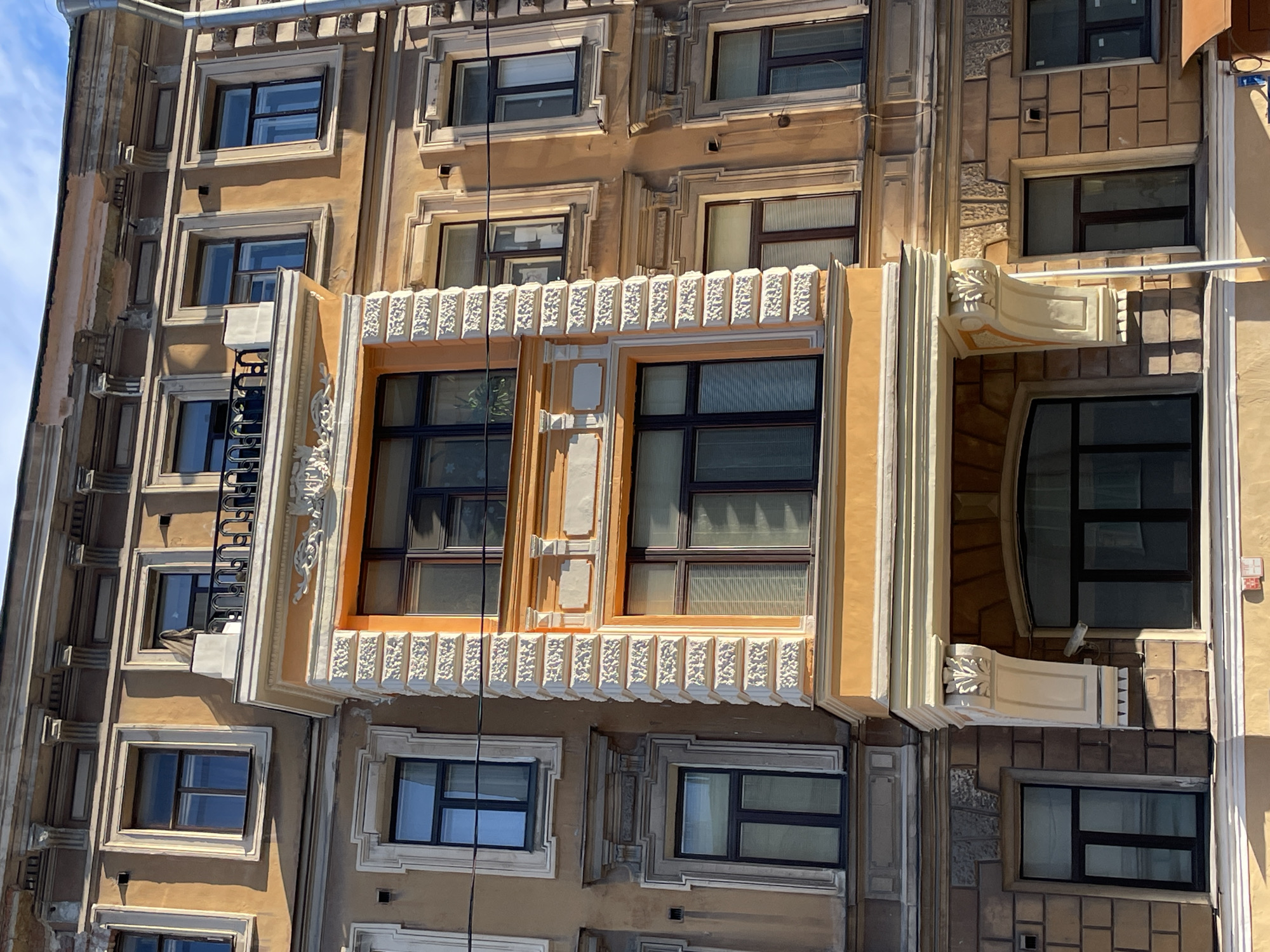 Exterior shot of a building in Saint Petersburg, Russia, with multiple windows of varying sizes, framed with decorative moldings. The building is light brown with white and cream architectural embellishments. Some windows have window panes, while others appear to be open. There is a protruding architectural feature that spans two windows on the middle floor, elaborately decorated. Utility cables are visible across the facade.