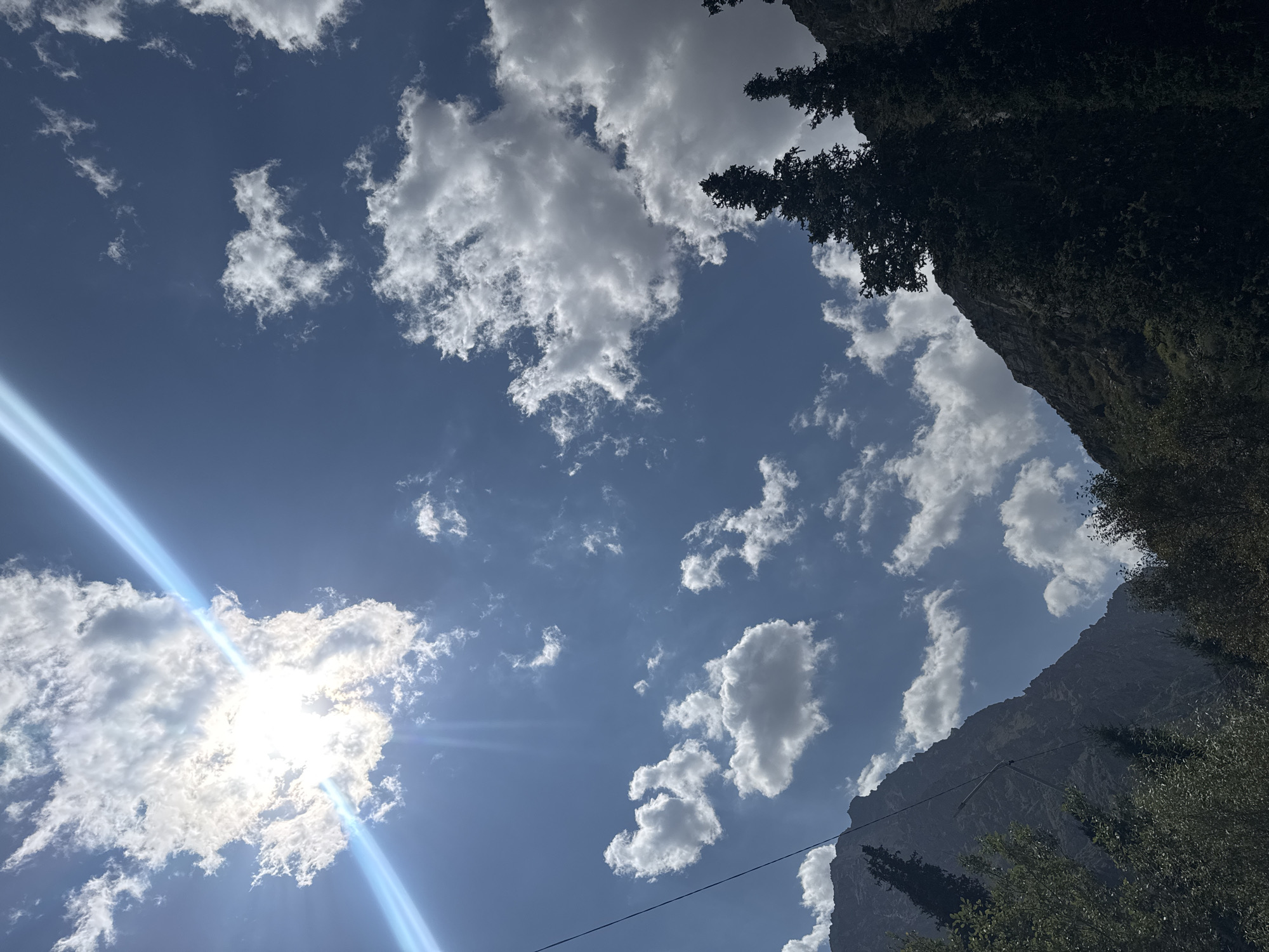 The image captures a dramatic sky over a mountainous landscape. The sun, partially obscured by a large cloud, casts a bright glare with visible light rays. Smaller, white cumulus clouds are scattered across a deep blue sky. In the foreground, the dark silhouettes of tall evergreen trees and the rugged slopes of mountains dominate the lower portion of the frame. A thin black wire, possibly a power line or telephone wire, stretches diagonally across the left side of the image. The scene appears to be a natural, possibly remote, location in Kyrgyzstan, given the mountainous terrain and vegetation. No people or animals are visible.