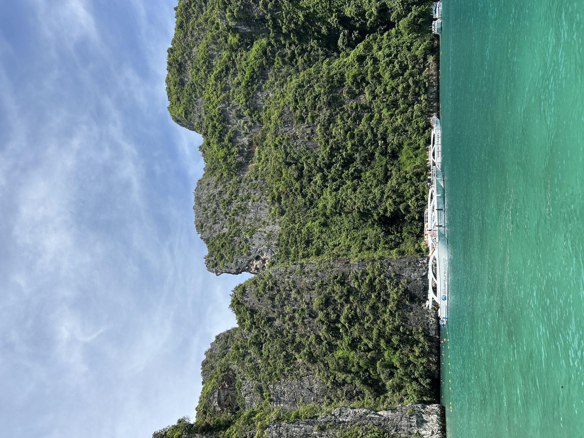A wide-angle, eye-level shot captures a serene bay in Ao Nang, Thailand, dominated by towering limestone karsts carpeted in lush green vegetation. The water in the foreground is a vibrant, clear turquoise, gently rippling under a bright blue sky dotted with wispy white clouds. Several white tour boats are moored in the bay, with passengers visible on deck. One boat prominently displays the text "adventureworld." A line of yellow buoys marks a swimming area, and a few people can be seen swimming and relaxing in the water. The karsts, with their rugged textures and dramatic vertical formations, create a natural amphitheater around the tranquil bay. The scene evokes a sense of tropical paradise and popular tourist activity.
