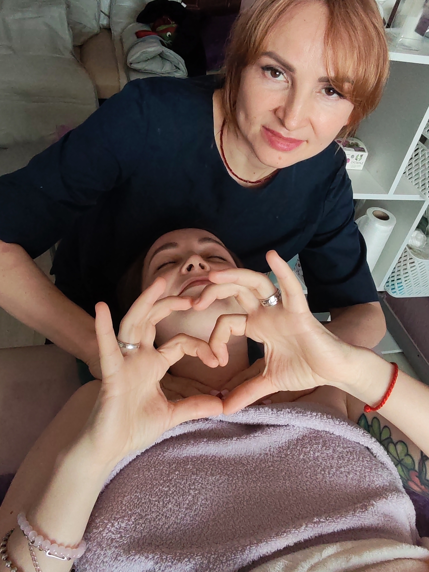 The image shows two people. A woman with reddish-brown hair and a black top is behind a person lying down. The person lying down has their eyes closed and is making a heart shape with their hands in front of their face. The person is covered with a light purple towel. The location is likely indoors, with shelves and a roll of paper visible in the background. There is a pillow and assorted items on a couch behind the woman standing. The exact location is Chaikovsky, Russia.