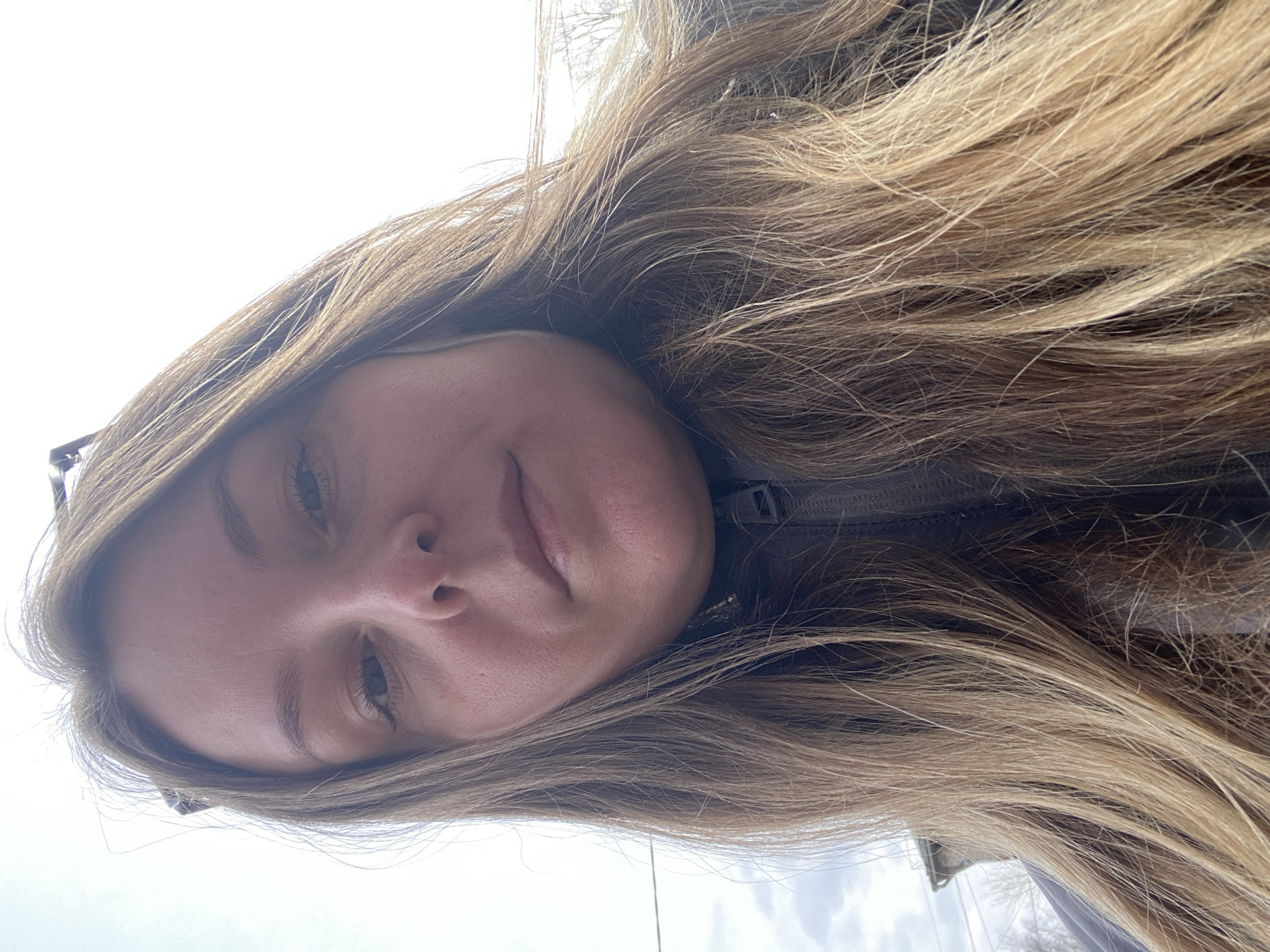 A selfie shot from a low angle depicts a person with long, light brown hair and fair skin. The background shows a cloudy sky and what appears to be a blurred outdoor scene with trees, likely taken in daylight. The person is wearing a dark-colored jacket. There is no explicit evidence to confirm a location in Chaikovsky, Russia.