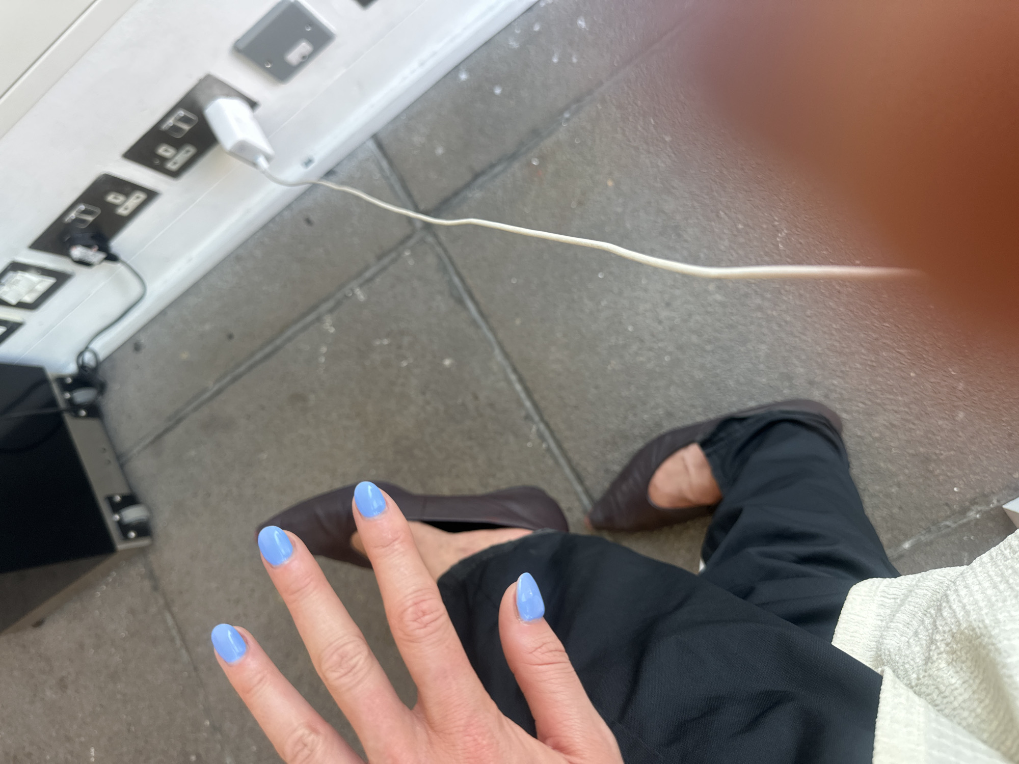 A first-person perspective photograph shows a person sitting with their feet stretched out in front of them, apparently in a public area where charging outlets are available. The individual has their hand extended towards the camera, showing off their painted, light blue fingernails. They are wearing dark-colored pants and brown ballet flats. A white charging cable extends from a wall-mounted power outlet with multiple sockets, some of which are occupied. The foreground and background surface appears to be tiled or paved, typical of urban walkways. Based on the plug types, this could be in the United Kingdom. Given the presence of public charging stations, it's likely a busy transit area, such as an airport, train station, or shopping center in the city of London.