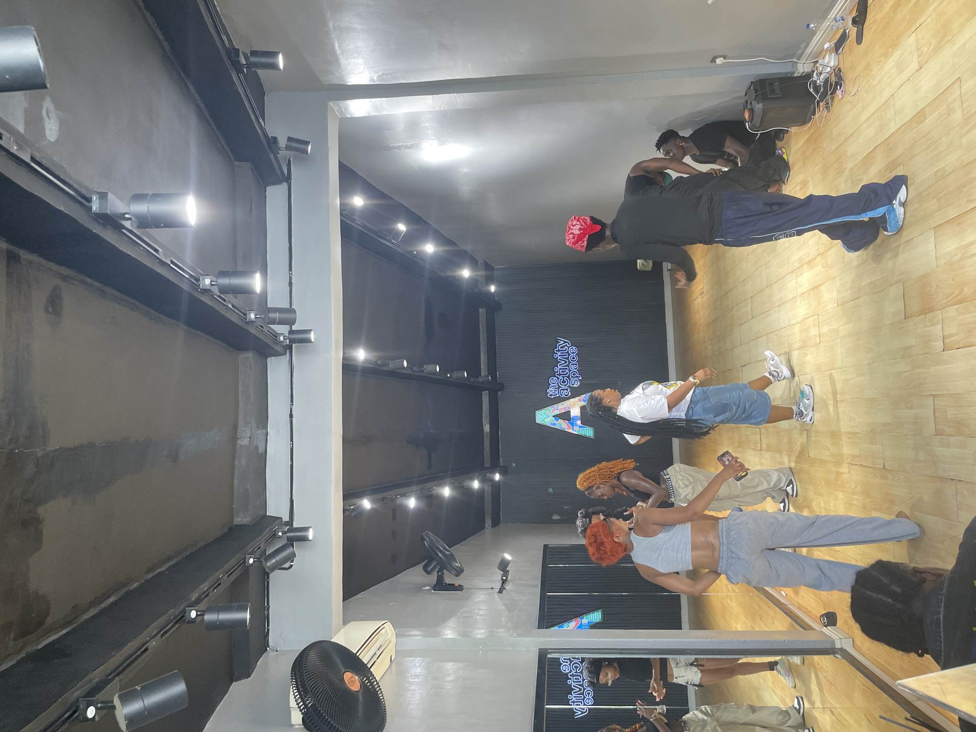 The image shows a group of individuals in a dance studio located in Lekki, Nigeria. The studio features a wooden floor and mirrored walls. Several individuals are standing and interacting, while one person is lying on the floor. The studio is equipped with track lighting and a large speaker system. A logo of "The Activity Space" is visible on one of the walls. Fans are present on the floor.
