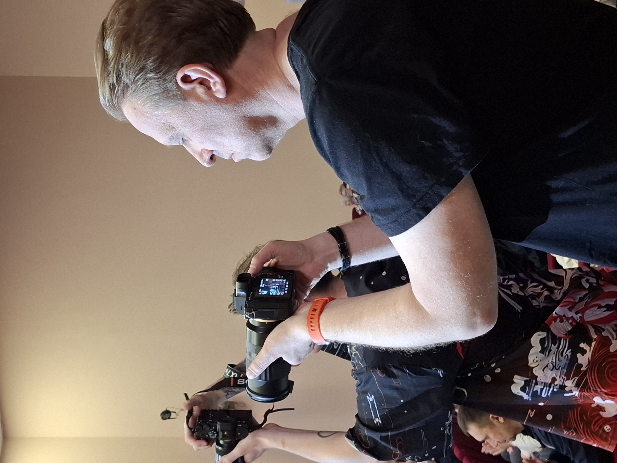 A medium shot shows three people, two of whom are holding cameras. The man in the foreground is fair-skinned and is wearing a black t-shirt. He is looking down at the camera held in his hands. The man behind him is wearing a patterned shirt and holding a camera, with an orange wristband on his wrist. Another person can be seen behind him. The background is a beige wall. The image was taken in a location that is not possible to establish from the context of the image.