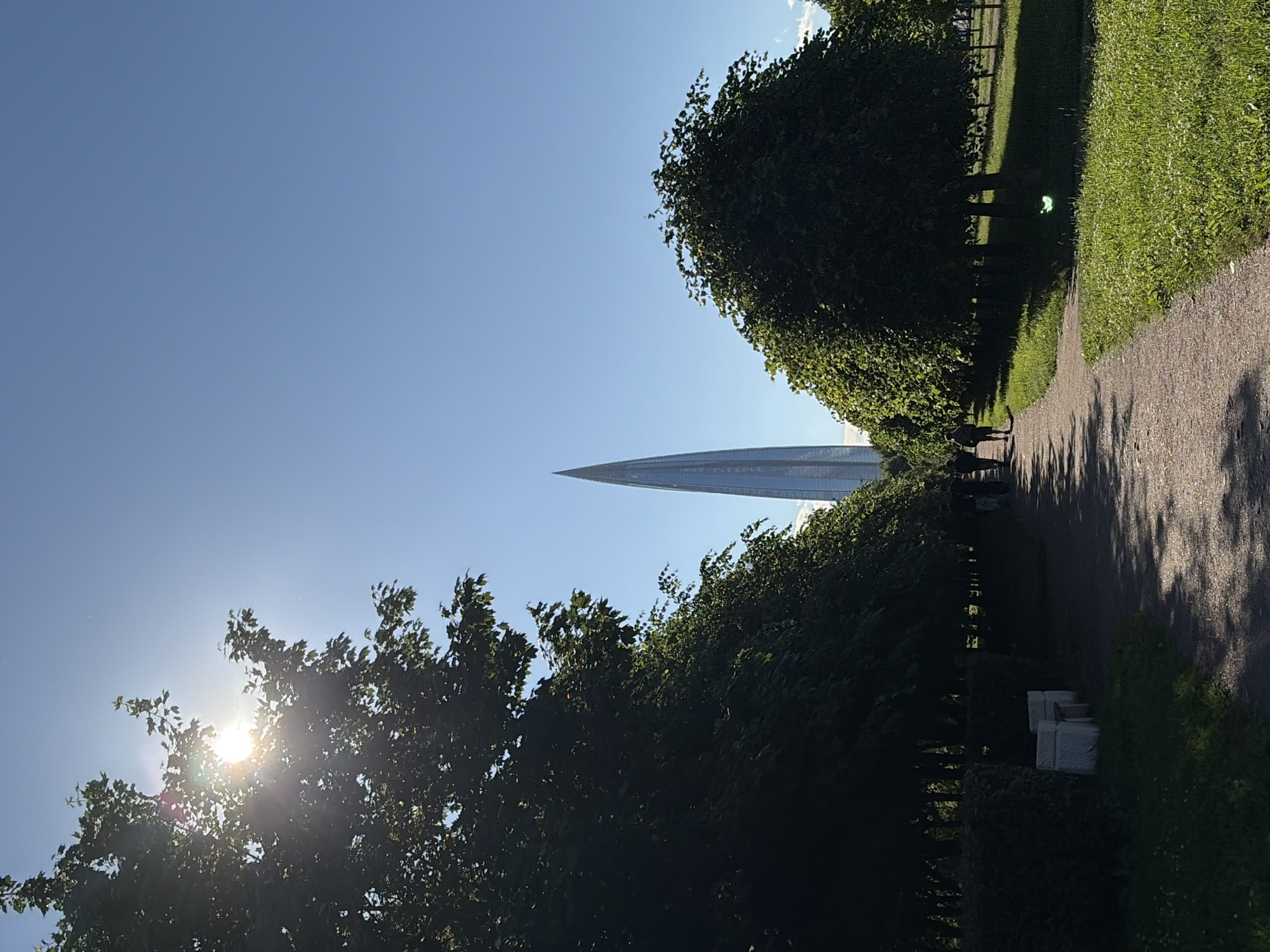 The image captures a sun-drenched park in Saint Petersburg, Russia, with the distinctive Lakhta Center skyscraper prominently visible in the distance. The foreground features a paved pathway, bordered by lush green grass and mature trees with dense foliage. Sunlight filters through the leaves of a large tree on the left, creating dappled light patterns on the path and grass. Several figures, appearing to be pedestrians, are seen walking along the path in the middle ground, silhouetted against the bright sky. On the left side of the path, there are two white park benches partially obscured by shadows. The Lakhta Center, a striking glass-clad skyscraper with a pointed spire, dominates the background, standing tall against a clear blue sky. The overall scene conveys a peaceful and sunny day in an urban park.