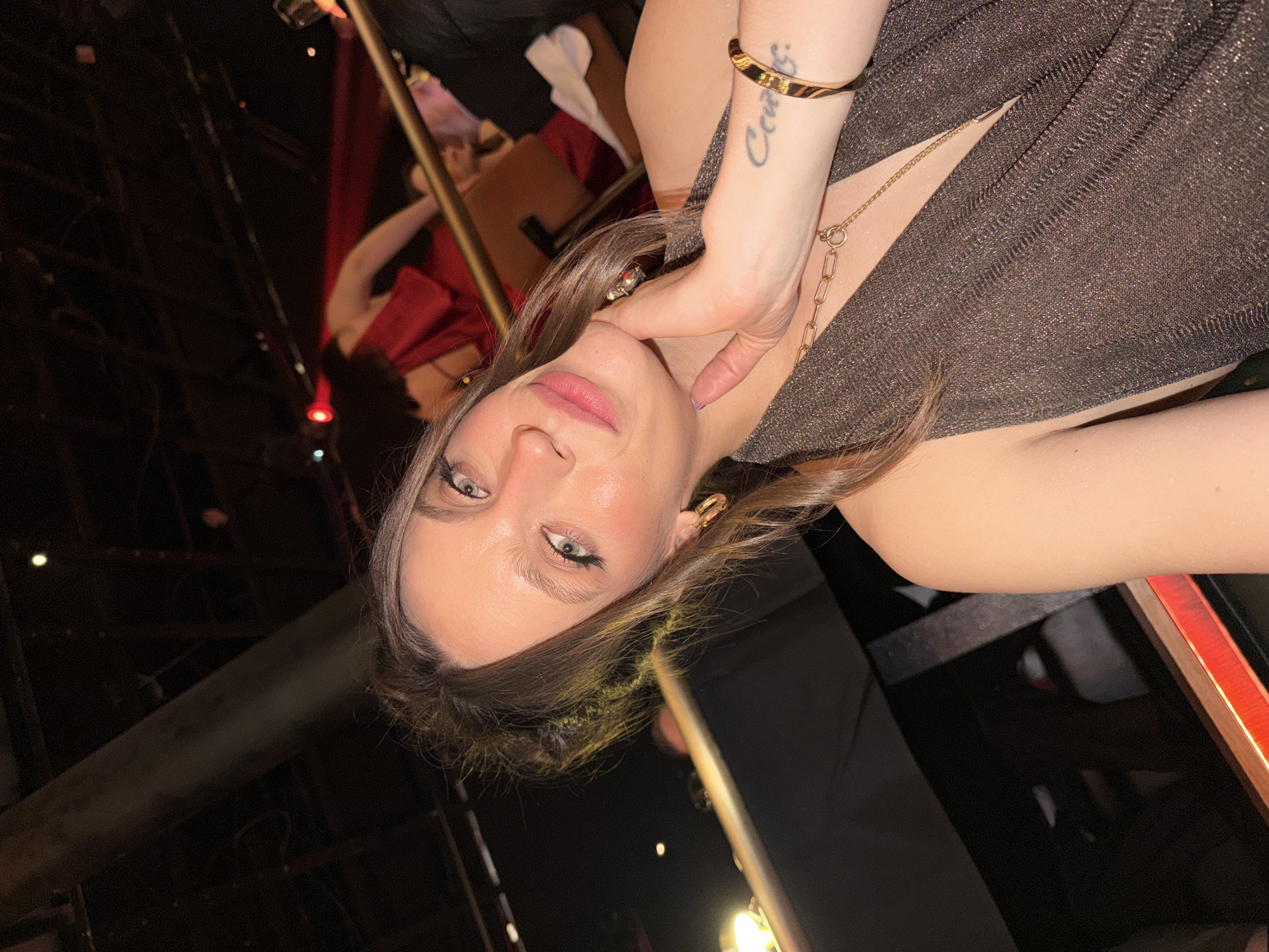 A woman is posing for a photograph indoors. The woman has light skin, hazel eyes, and light brown hair, and is wearing a dark brown, sparkling dress and gold earrings and a gold chain necklace. She is touching her neck with her right hand, which also has a gold bracelet on the wrist. A tattoo is visible on her forearm. Behind her, there are dark, blurred figures and furniture with red accents, suggesting a bar or club setting. The location is likely Belgrade, Serbia.