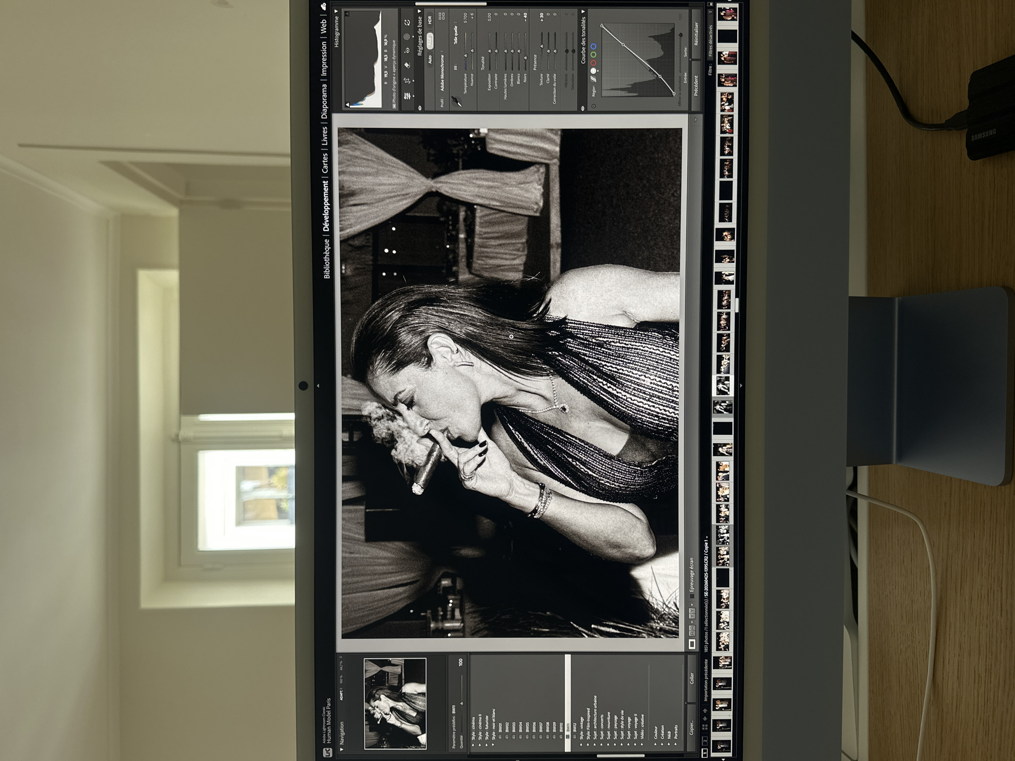 The primary image displays a computer monitor showing the Adobe Lightroom Classic editing software, rotated 90 degrees clockwise. The software's interface is in French and presents a black and white photograph of a woman.
The woman in the photograph has long, dark hair and is wearing a shimmering or sequined top. She is actively smoking a cigar, holding it between her fingers. Rings are visible on her fingers and bracelets on her wrist. Her expression appears focused or contemplative, with her eyes partially closed. The background of her image is dark and suggests an indoor setting.
On the monitor, the Lightroom interface shows the "Développement" (Develop) module. Visible panels include "Réglages de base" (Basic Adjustments) with sliders for exposure, contrast, highlights, shadows, whites, blacks, texture, clarity, and dehaze. The "Courbe des tonalités" (Tone Curve) is also displayed. The image is being processed in black and white, as indicated by the "Paramètre prédéfini: BW" (Preset: B&W) section. A navigation panel at the bottom left includes the text "Adobe Lightroom Classic" and "Human Model Paris," implying the photo's context is in Paris, France.
The real-world environment surrounding the monitor shows a bright room with light-colored walls and a window providing natural daylight. A wooden surface is visible on the right side of the monitor, where a white cable is connected to a black charger with the "SAMSUNG" logo.
