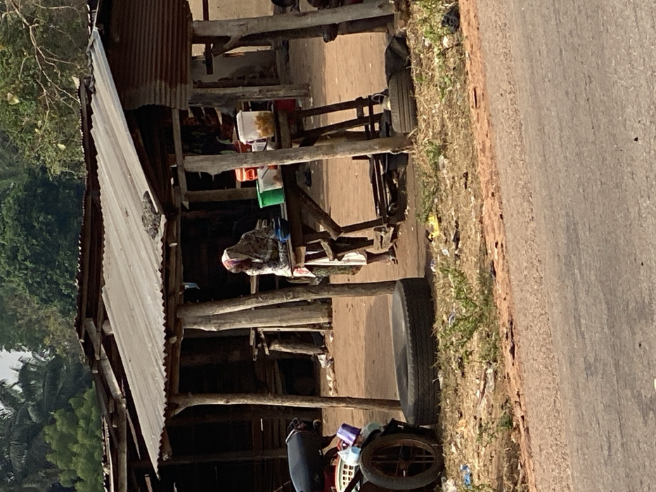 This image depicts a roadside scene in Okura, Nigeria. The foreground shows a paved road surface adjacent to an unpaved, reddish-brown dirt area. This dirt area contains sparse dry vegetation and scattered refuse, including plastic bags and debris. A single black vehicle tire lies flat on the ground in the mid-left.
At the center, a makeshift stall or shop structure is visible, featuring a weathered corrugated metal roof supported by multiple rough-hewn wooden poles. Under this structure, one individual, possibly a woman, stands or leans behind a wooden table. Her head is covered with a patterned fabric, and she wears a light-colored, long garment. Her face is obscured. On the wooden table in front of her are several containers: a blue bowl, a green bucket, a white bucket, a reddish-orange container, and a translucent container holding numerous light-colored spherical items, likely food.
To the left of the stall, a portion of a dark-seated motorbike with a spoked wheel is visible, appearing to be parked. Behind the primary stall, other similar rustic structures with corrugated roofs and wooden supports are partially discernible. The background features dense green foliage and trees under what appears to be natural daylight.