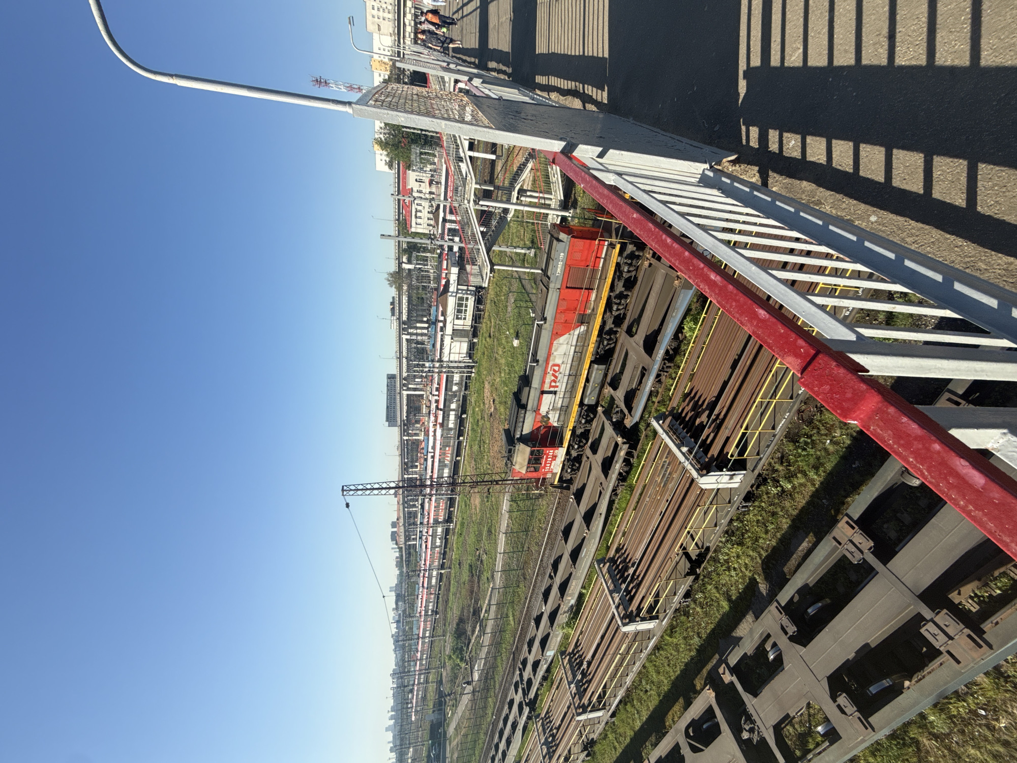 The image depicts a railway yard in Moscow, Russia, as viewed from an elevated pedestrian bridge. In the foreground, the bridge's metal railing is visible, casting shadows on the asphalt pathway. Beyond the railing, the railway yard stretches into the distance, filled with numerous train tracks, train cars, and a red and white locomotive with Cyrillic script. A construction crane is positioned in the middle ground, and in the far background, urban buildings can be seen. The sky is clear and blue. Several people are walking along the bridge.
